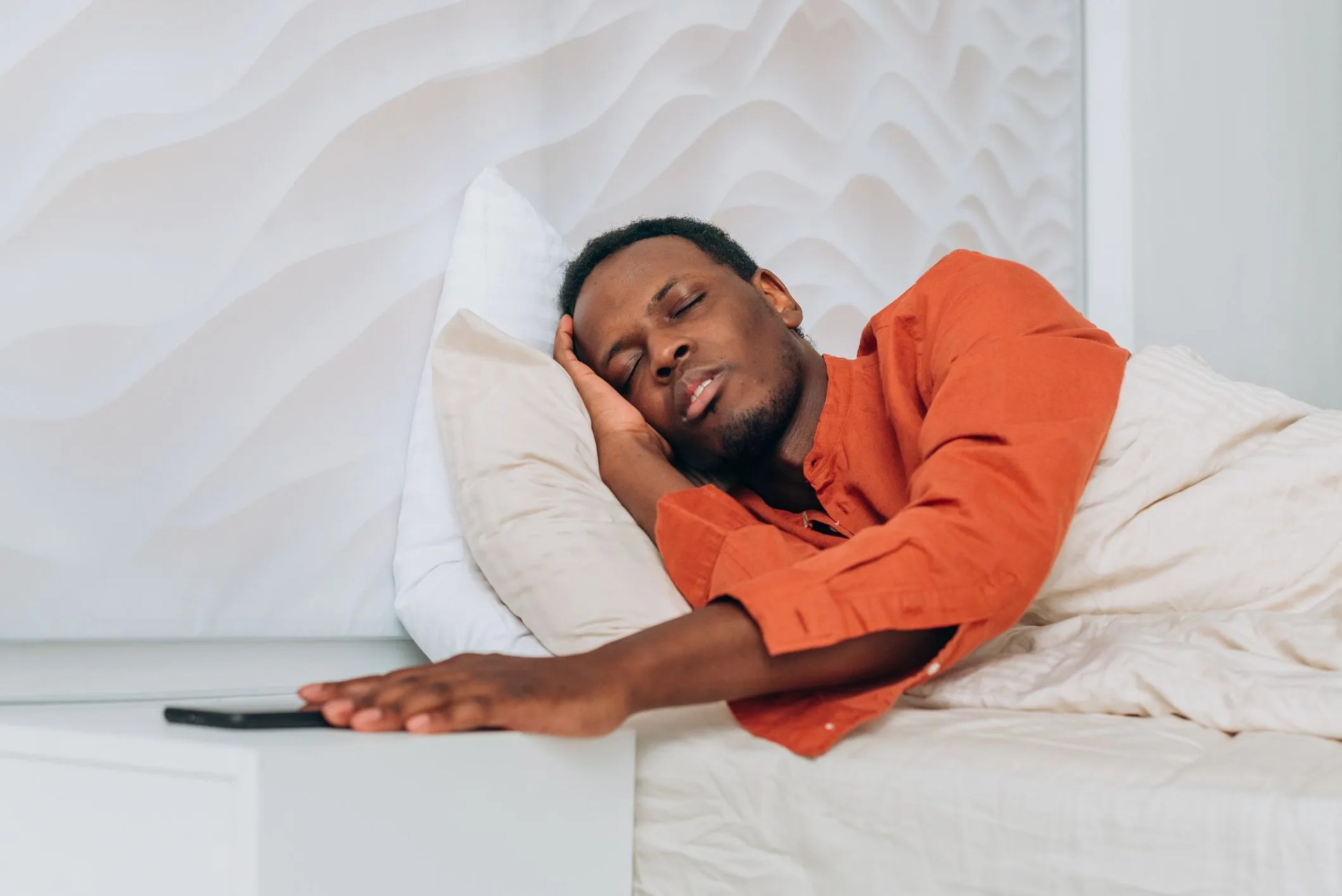 sleepy african american man in pajamas reaches to turn off alarm clock on smartphone awakening in large bed in room closeup