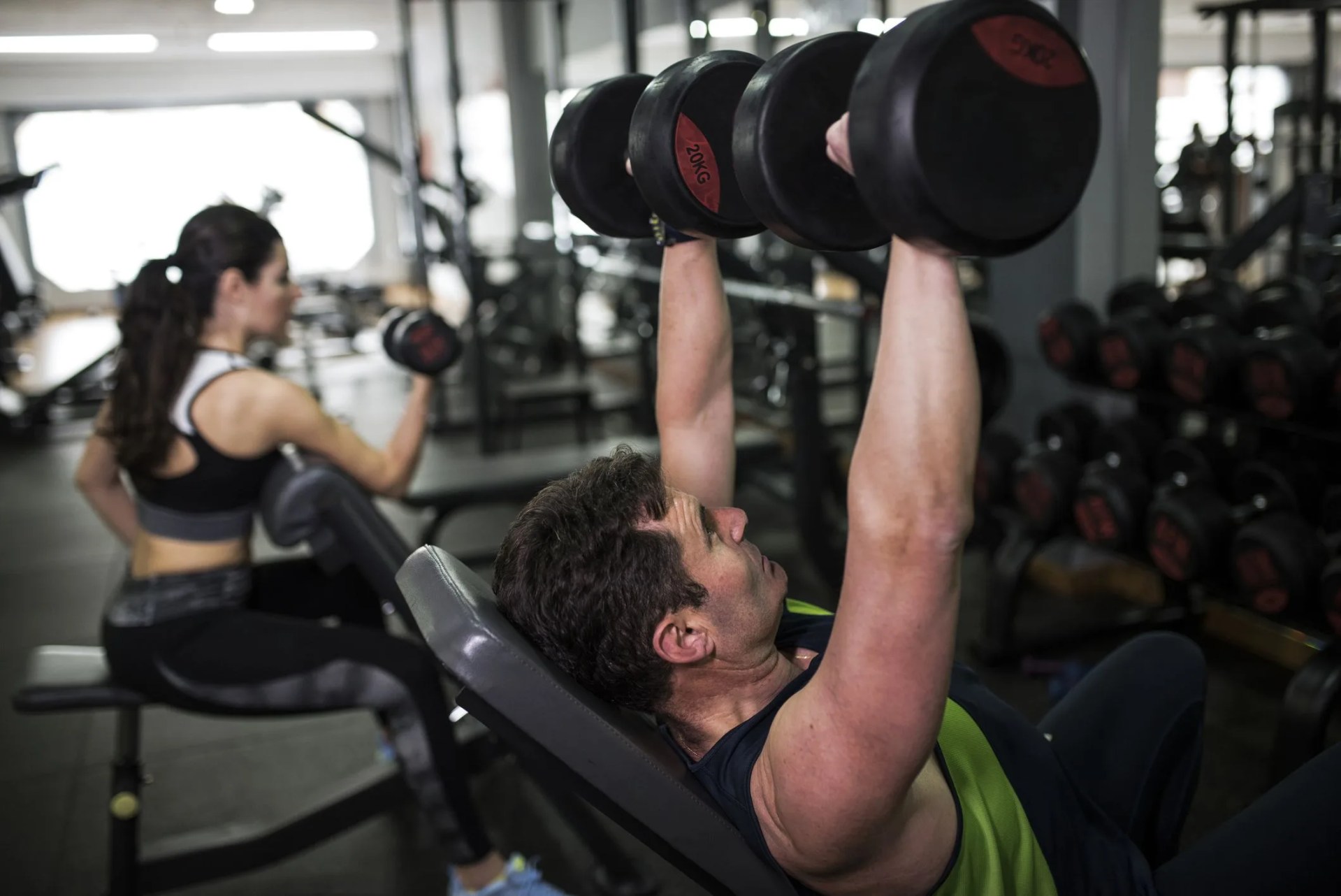 mature man in his 50s trains in the weights and dumbbell lifting to stimulate the chest in the gym to improve his strength performance 40 year old woman in the background, trains biceps