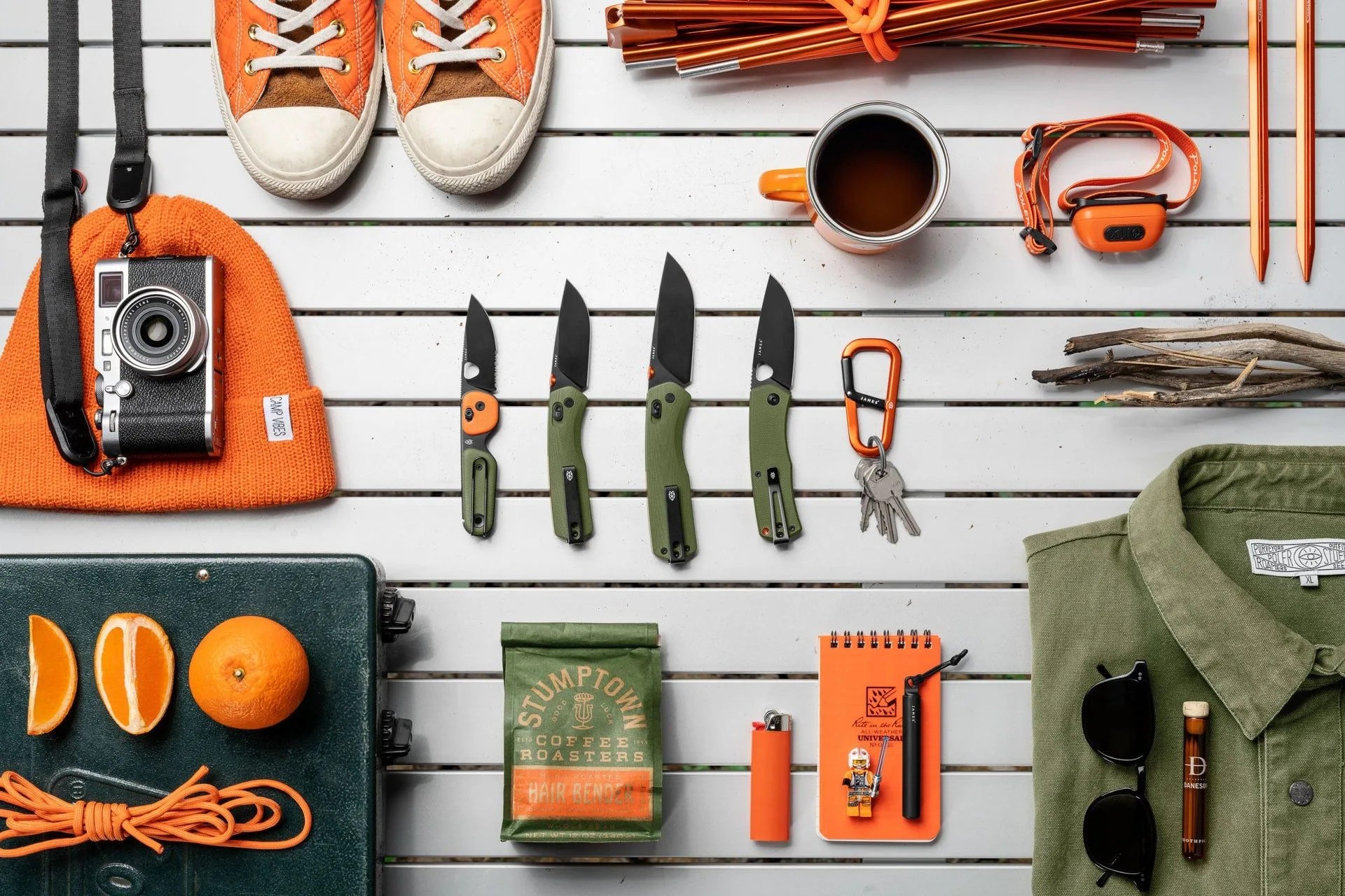 a flatlay photo of knives, a camera, camping gear, a shirt, sunglasses, and a cup of coffee