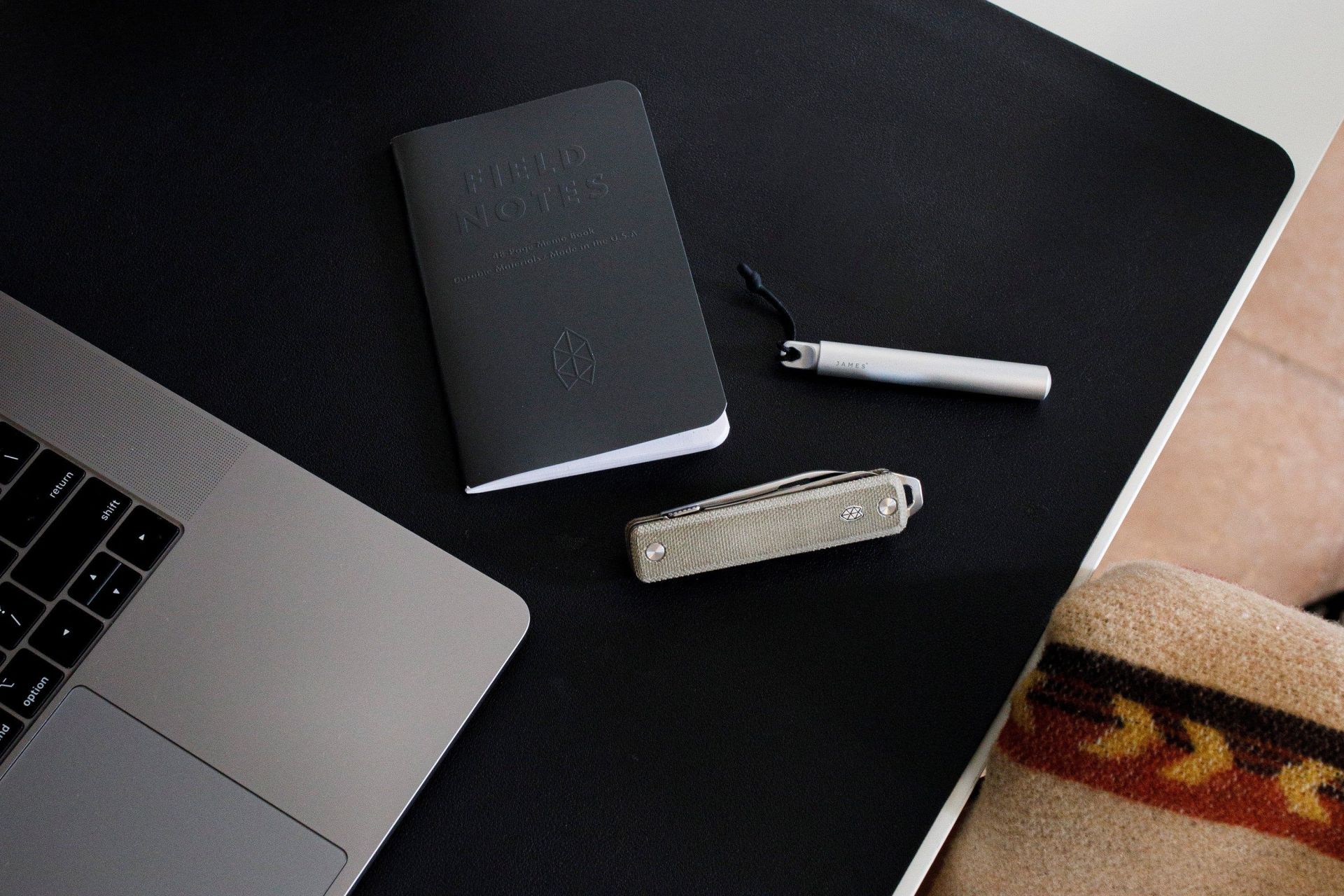 a notebook, pen, and pocket knife on a desk next to a laptop