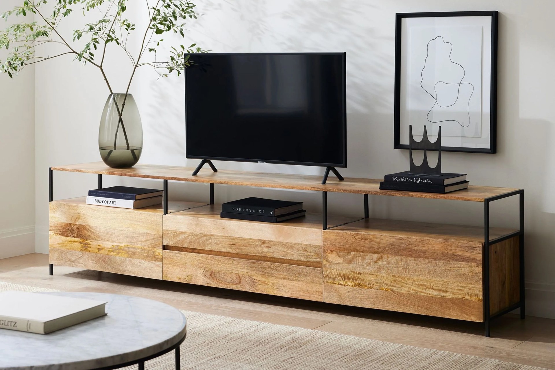 a tv on a wooden tv stand with a plant in a vase, art on the wall and books on a table