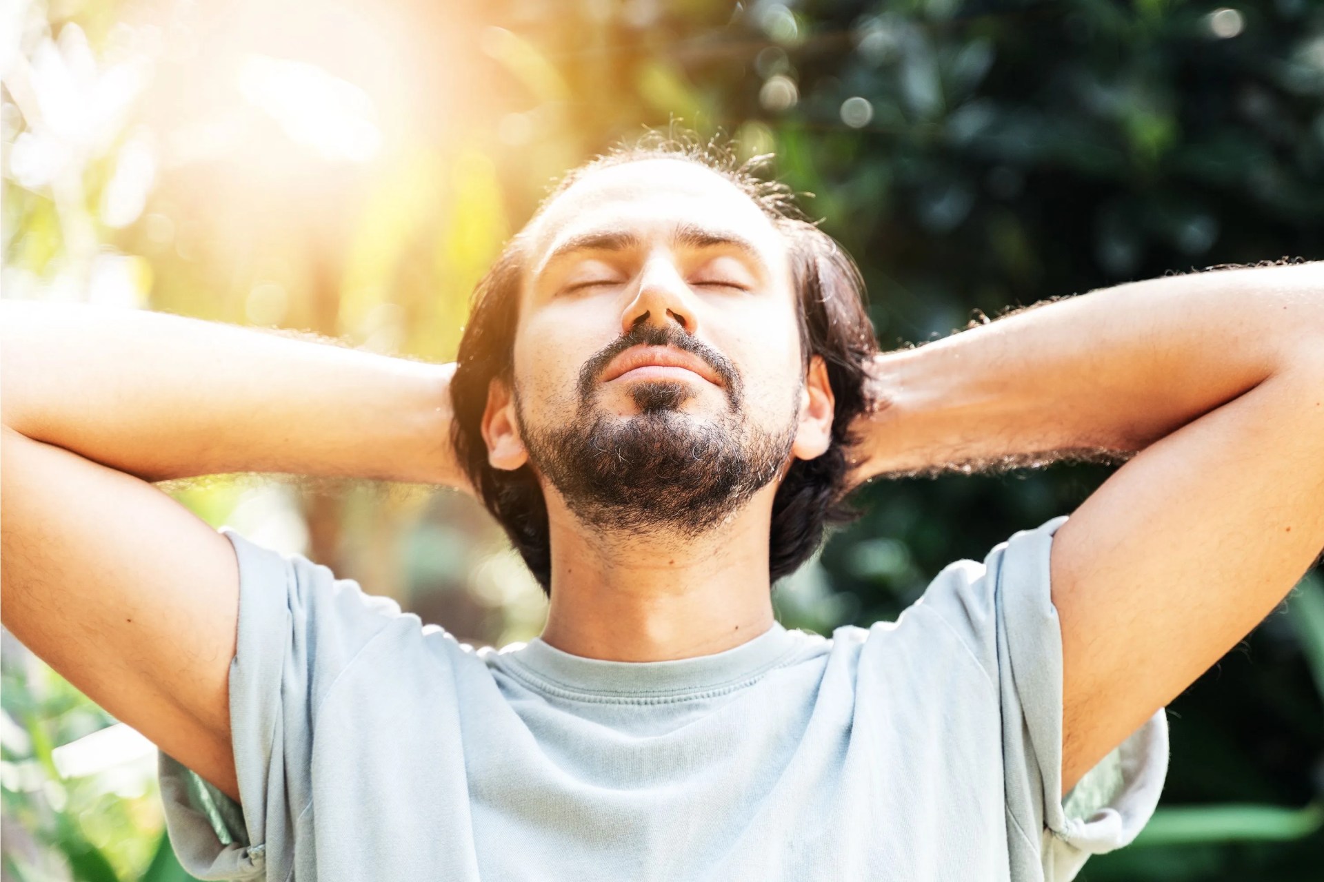 a bearded man is meditating outdoor in the park with face raised up to sky and eyes closed on sunny summer day