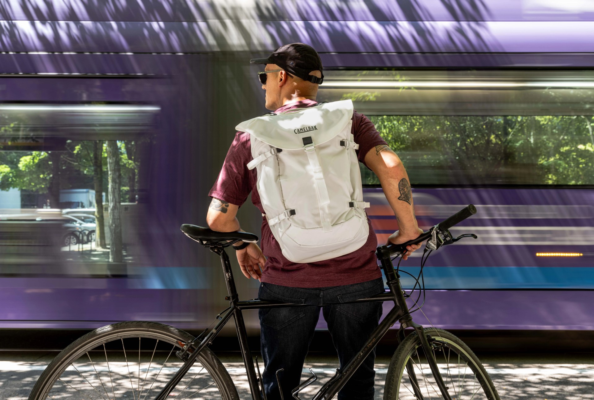 man standing at train station with bike as purple train goes by wearing camelbak backpack