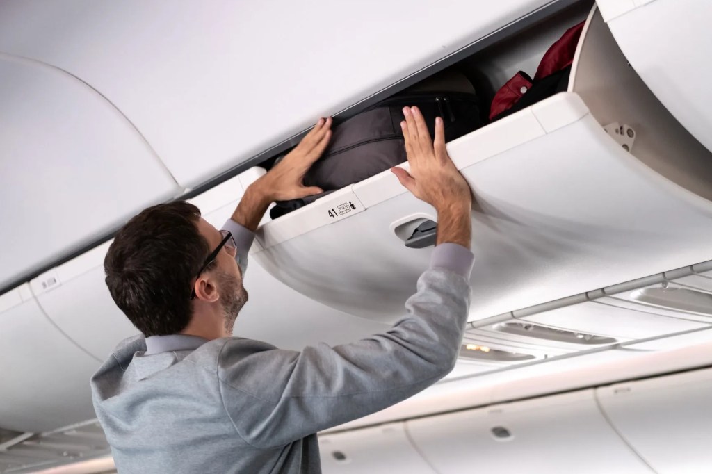 young man putting luggage into overhead locker on airplane traveler placing carry on bag in overhead compartment in aircraft