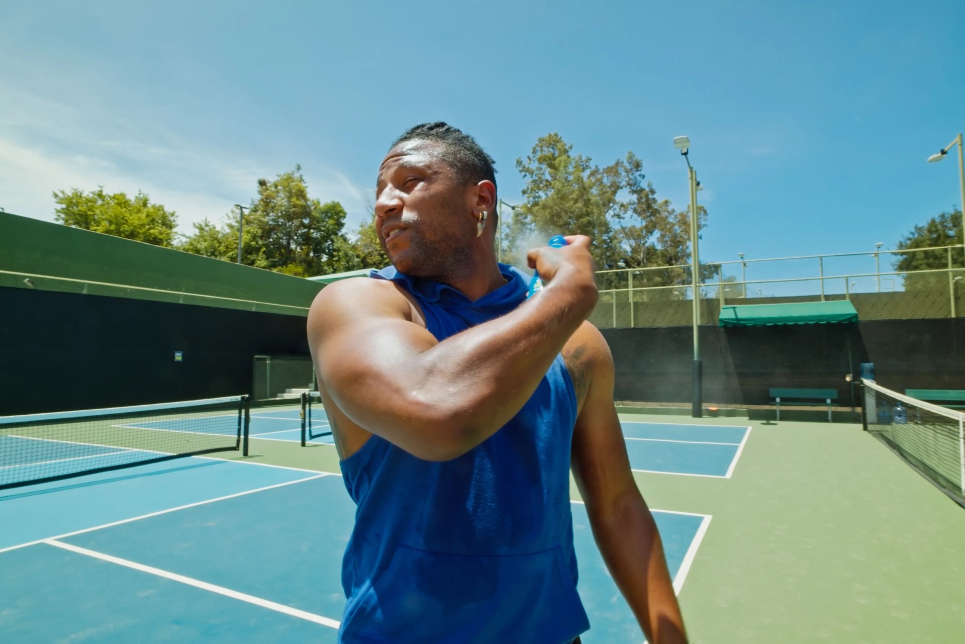 a young cape verdean man sprays on sunscreen on a pickleball court