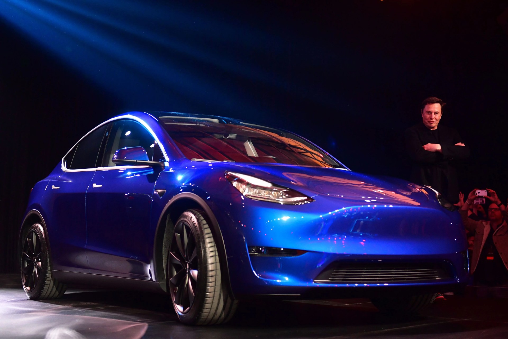 tesla model y on stage with elon musk in the background with arms folded staring at the camera