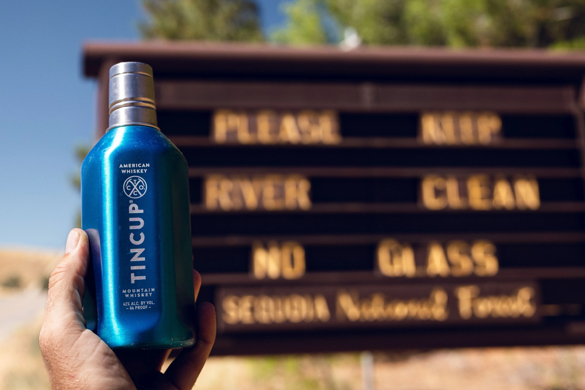 tincup whiskey held up against sign that reads please keep river clean no glass