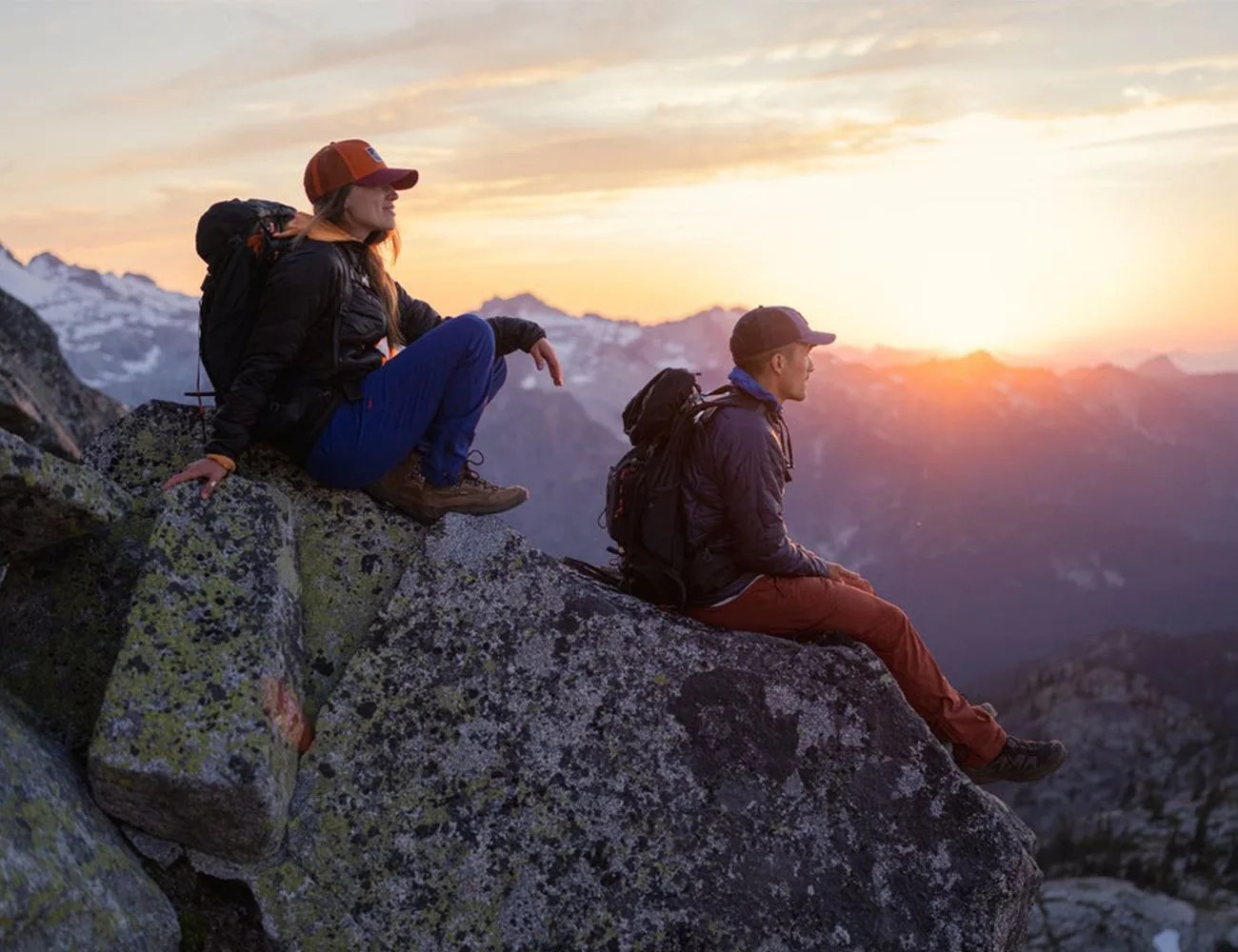two people sitting on a rocky mountain looking at the sun rise