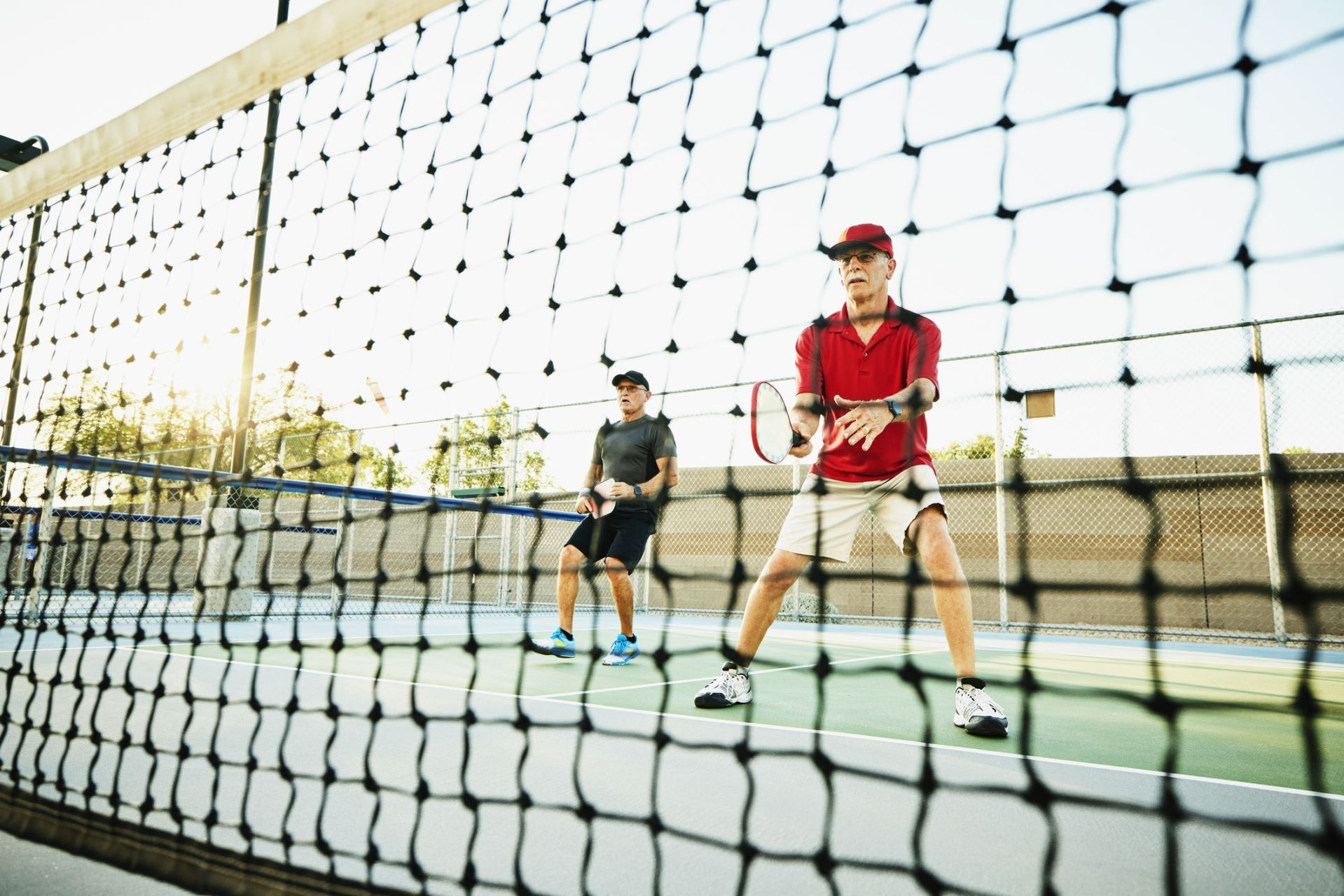 wide shot view through net of senior men playing doubles pickleball on summer evening