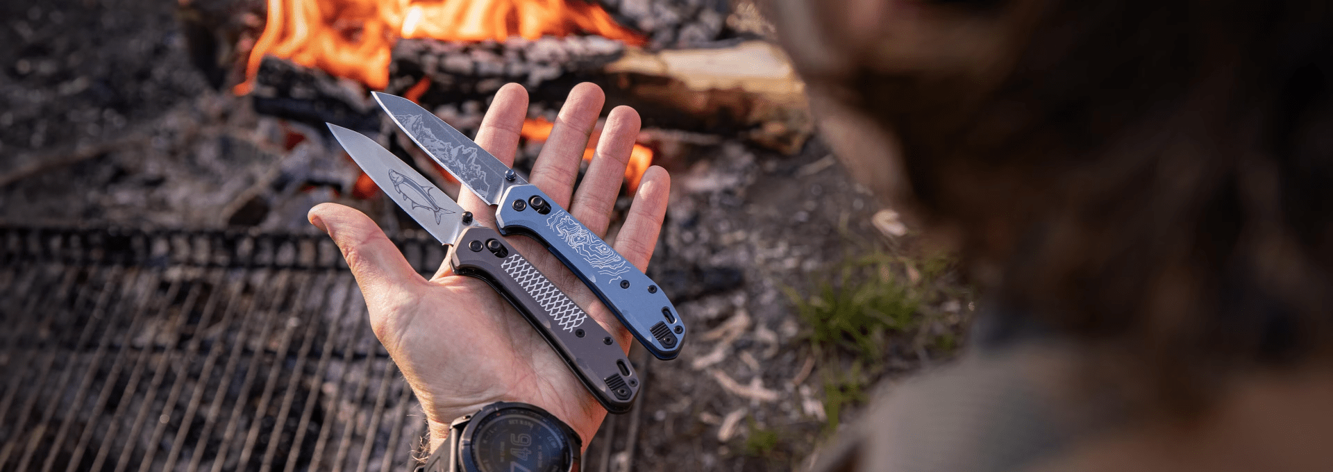 a hand holding two pocket knives in front of a fire
