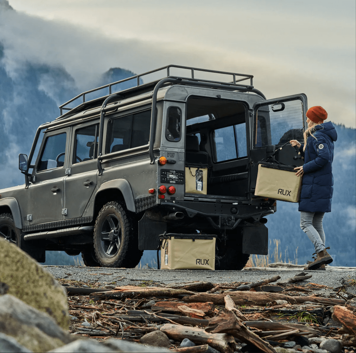 a woman holding a rux 70l packing it into the back of a land rover defender 110