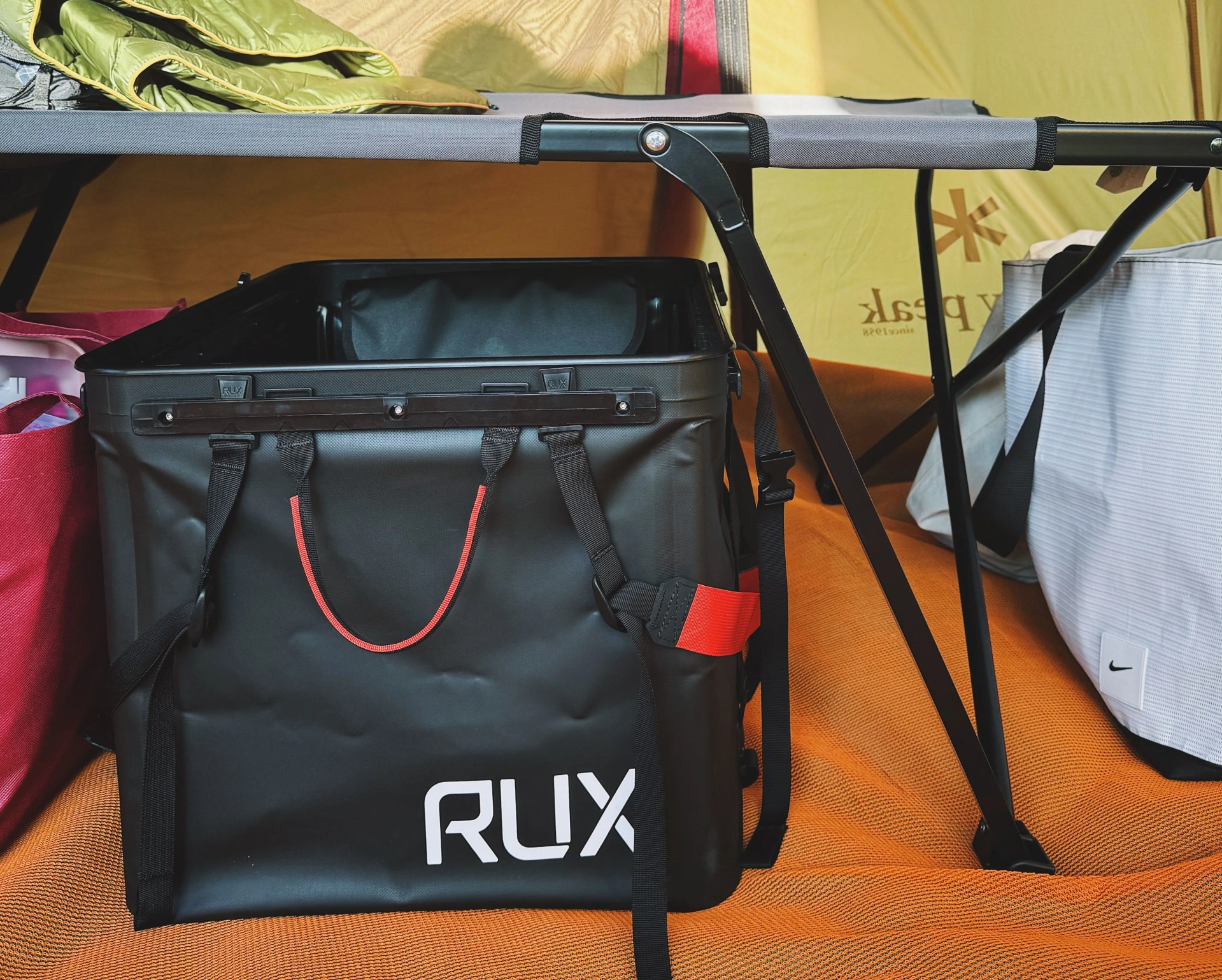 a black rux 70l stored under a camping cot