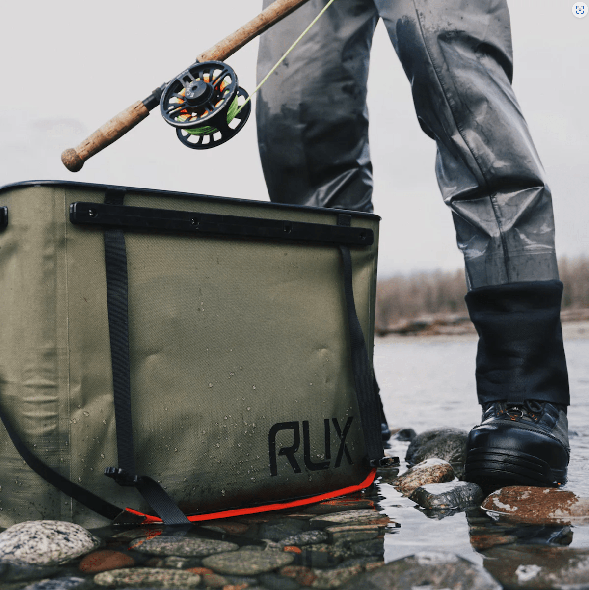 a green rux 70l bin sitting in a shallow river pool next to a person holding a fly fishing rod
