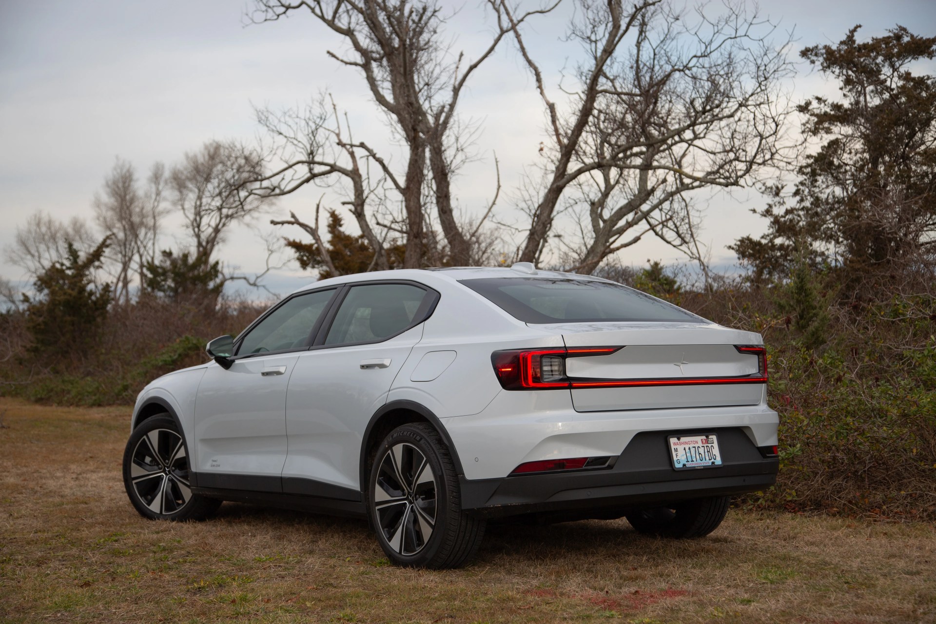 The rear three quarter angle of a white sedan parked on grass in front of brush and a tree with no leaves