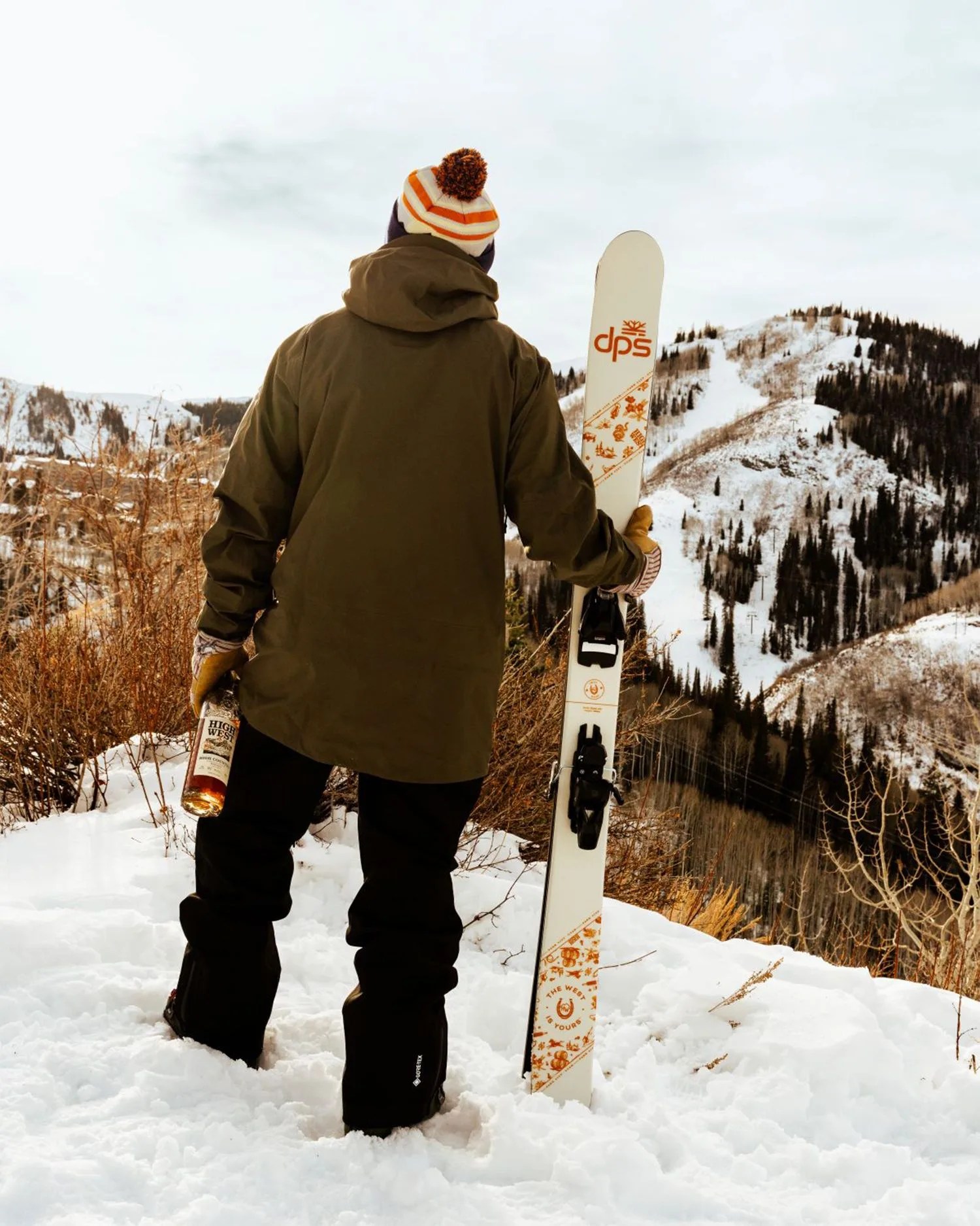 a man holding skiis and a bottle of whiskey on a snowy mountain