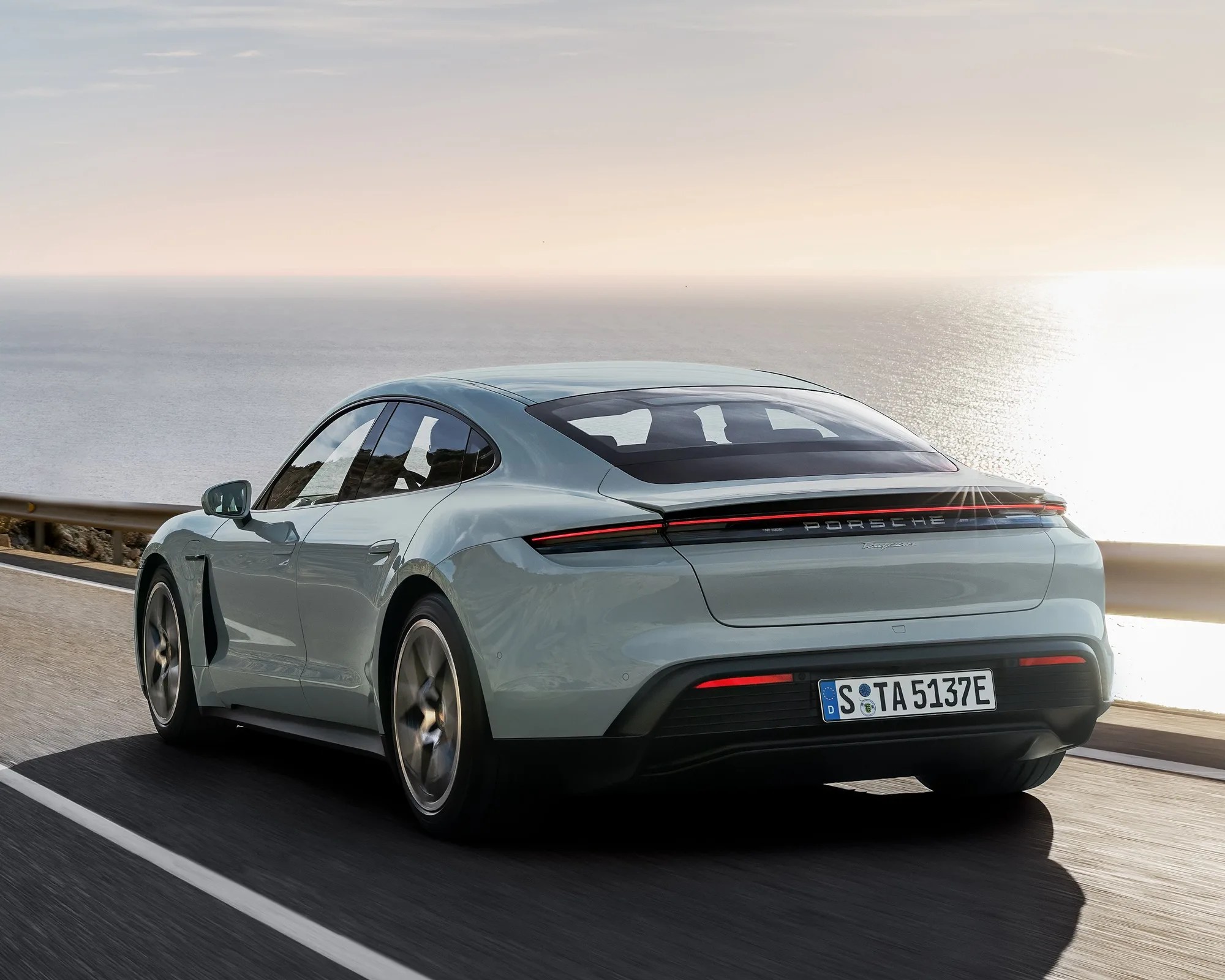 rear of light blue porsche taycan driving up a coastal road at sunset