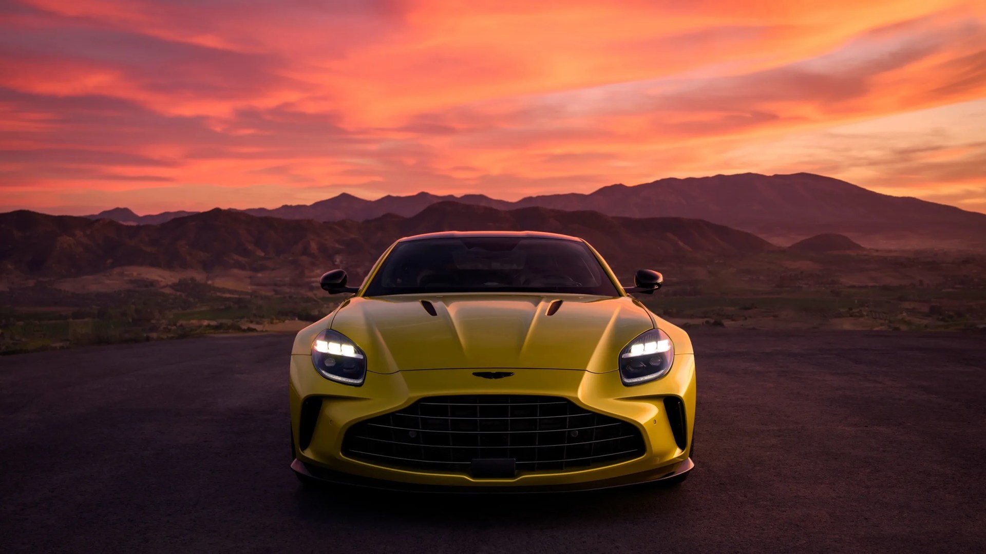 the front angle of a gold Aston Martin Vantage at sunset with purple mountains behind it in the distance