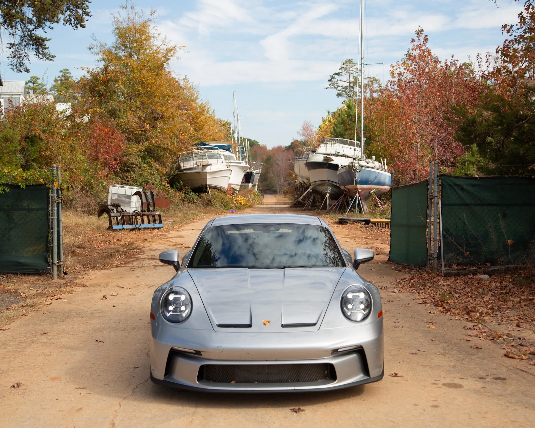 a silver porsche 911 gt3 touring parked facing you with boats on stands in the woods behind it
