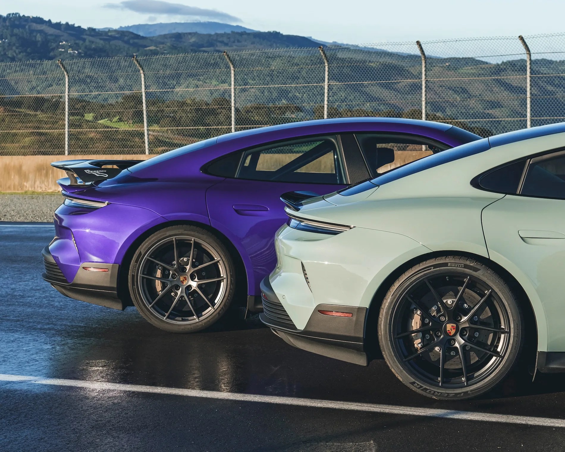 rear of a purple porsche taycan turbo gt and a green gray porsche taycan turbo gt parked next to each other on a race track with green hills in the back ground