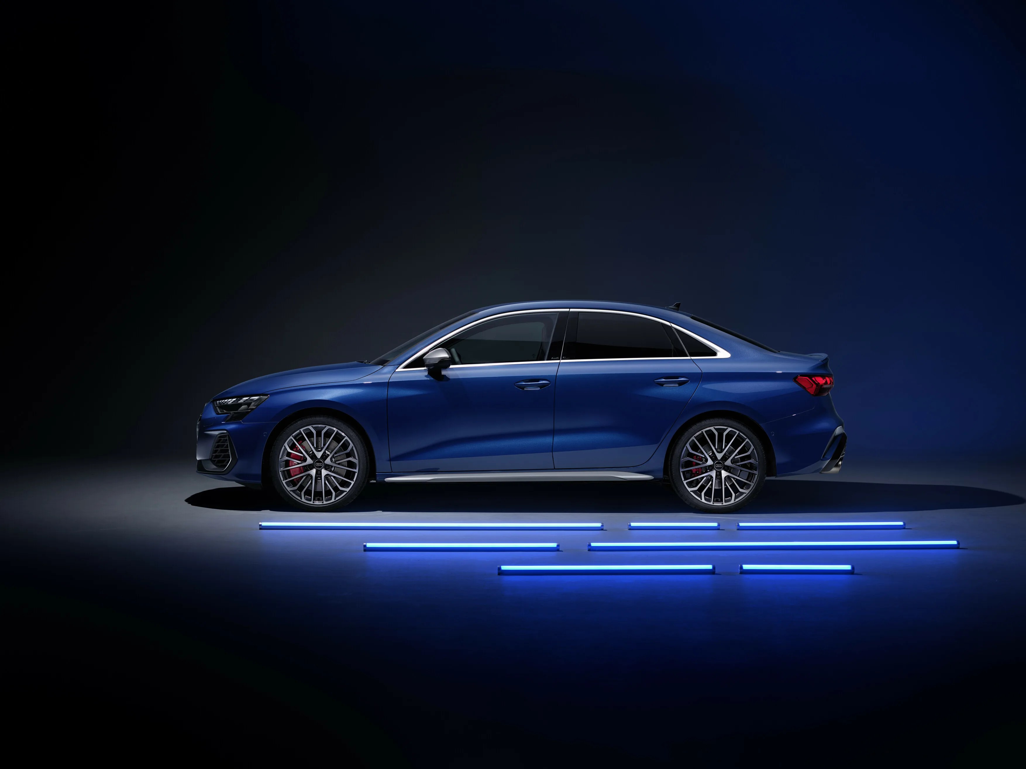 Blue 2025 Audi S3 sedan shown in profile next to blue neon lights on ground.