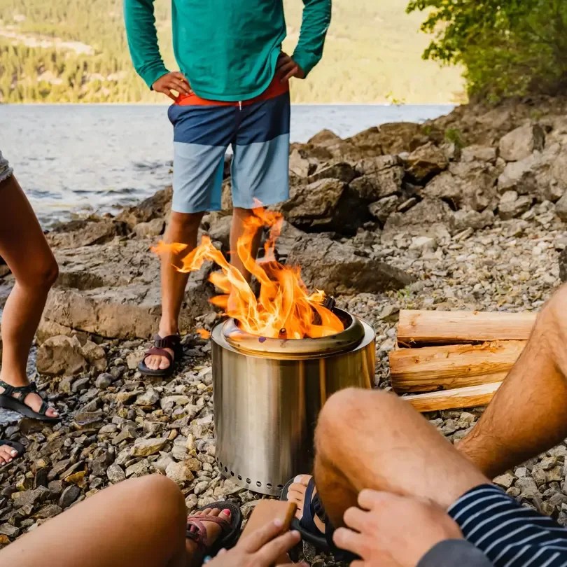 Group of people sitting and standing around a Solo Stove ranger on a rocky lake side shore with spare wood sitting on the ground. 