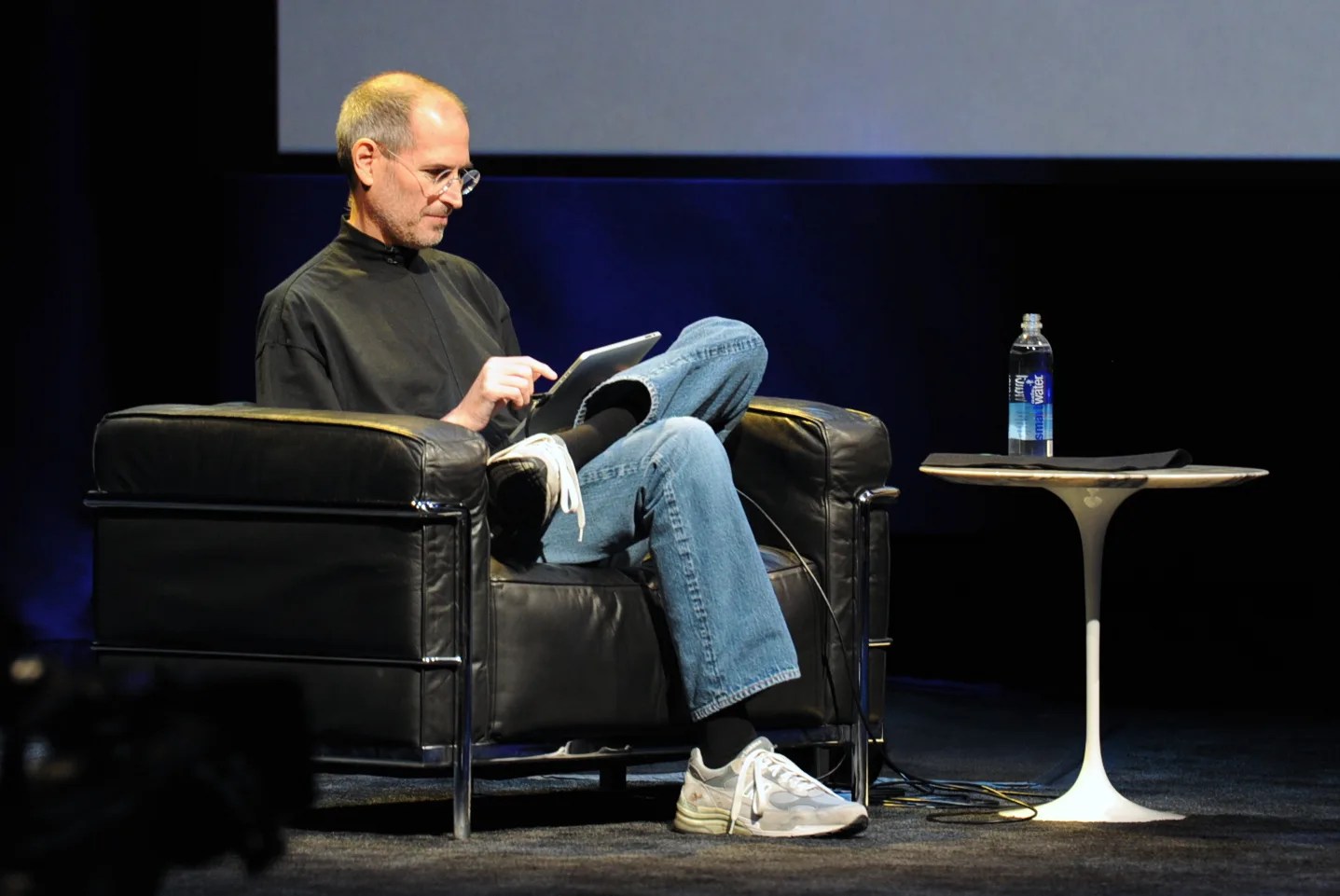 Steve Jobs sitting in black chair during original iPad presentation tapping on an iPad in his lap. 
