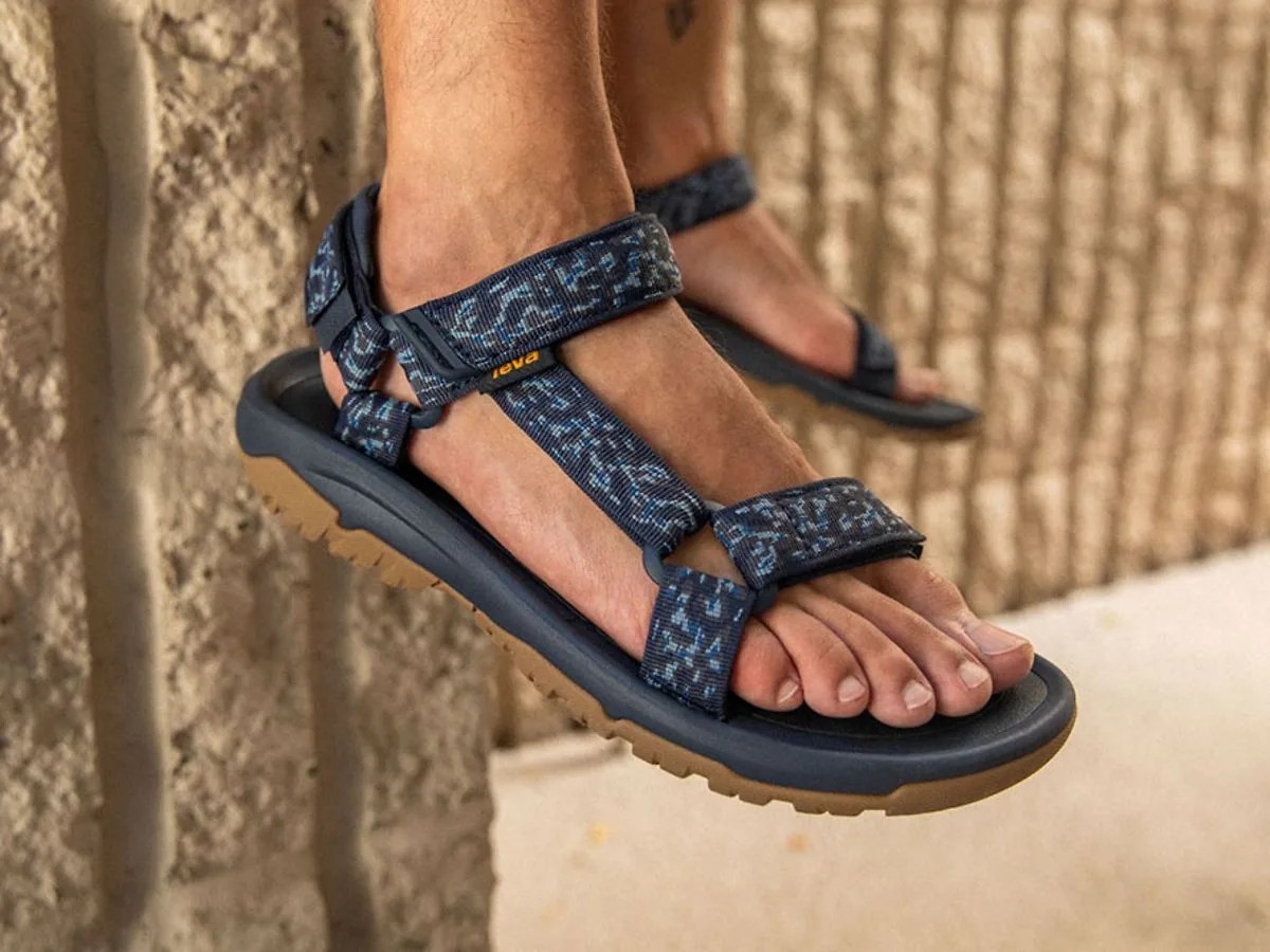 Teva Hurricane XLT on feet dangling in the air against a sand colored stone wall.