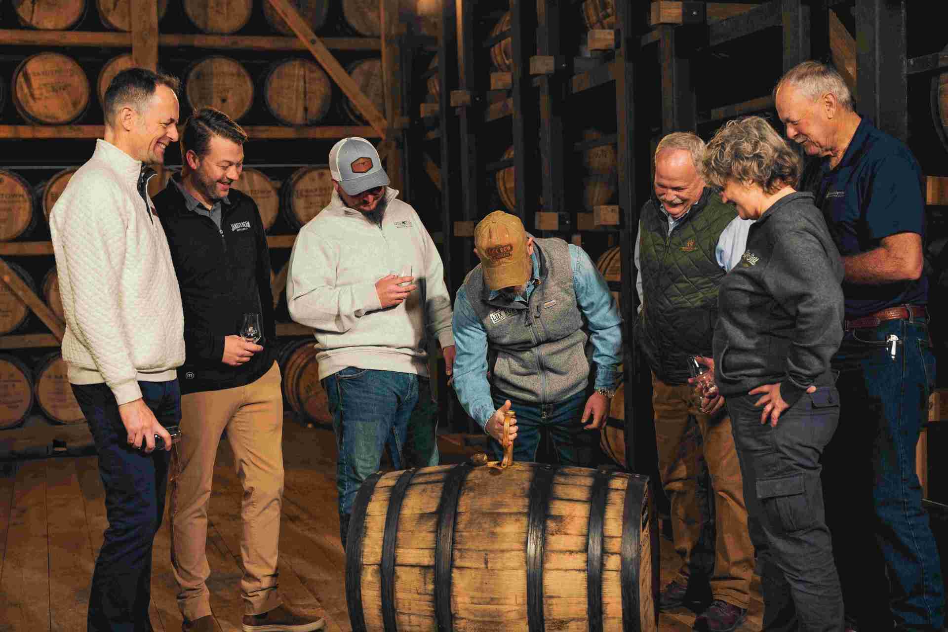 six men and one woman gather around a bourbon barrel