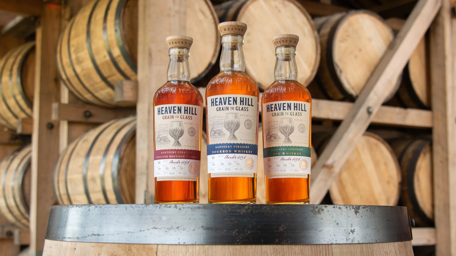 The three bottles of Heaven Hill’s Grain to Glass Whiskey Collection sitting on top of a bourbon barrel inside a bourbon barrel storage warehouse