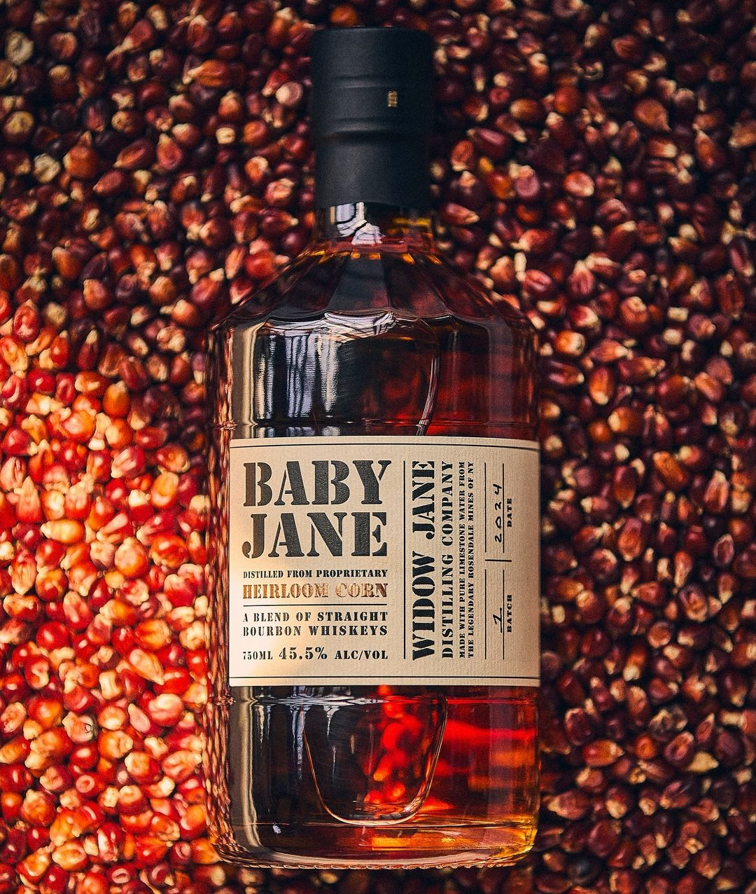 A bottle of Baby Jane bourbon sitting flat on a bed of red dried Baby Jane corn kernels.