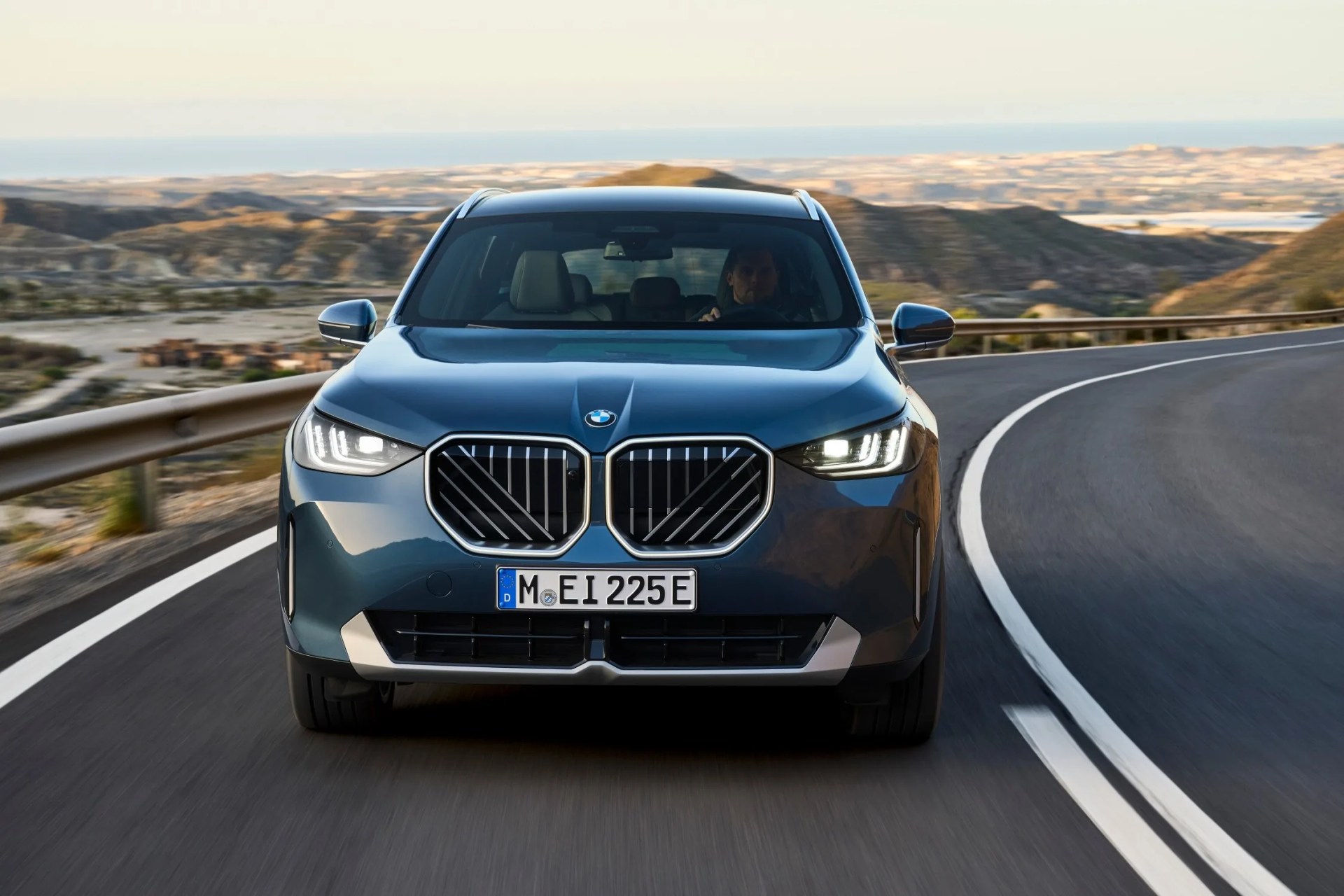 2025 BMW X3 in blue shown head on driving down the road highlighting the new models grill which features both vertical and diagonal lines for the first time.