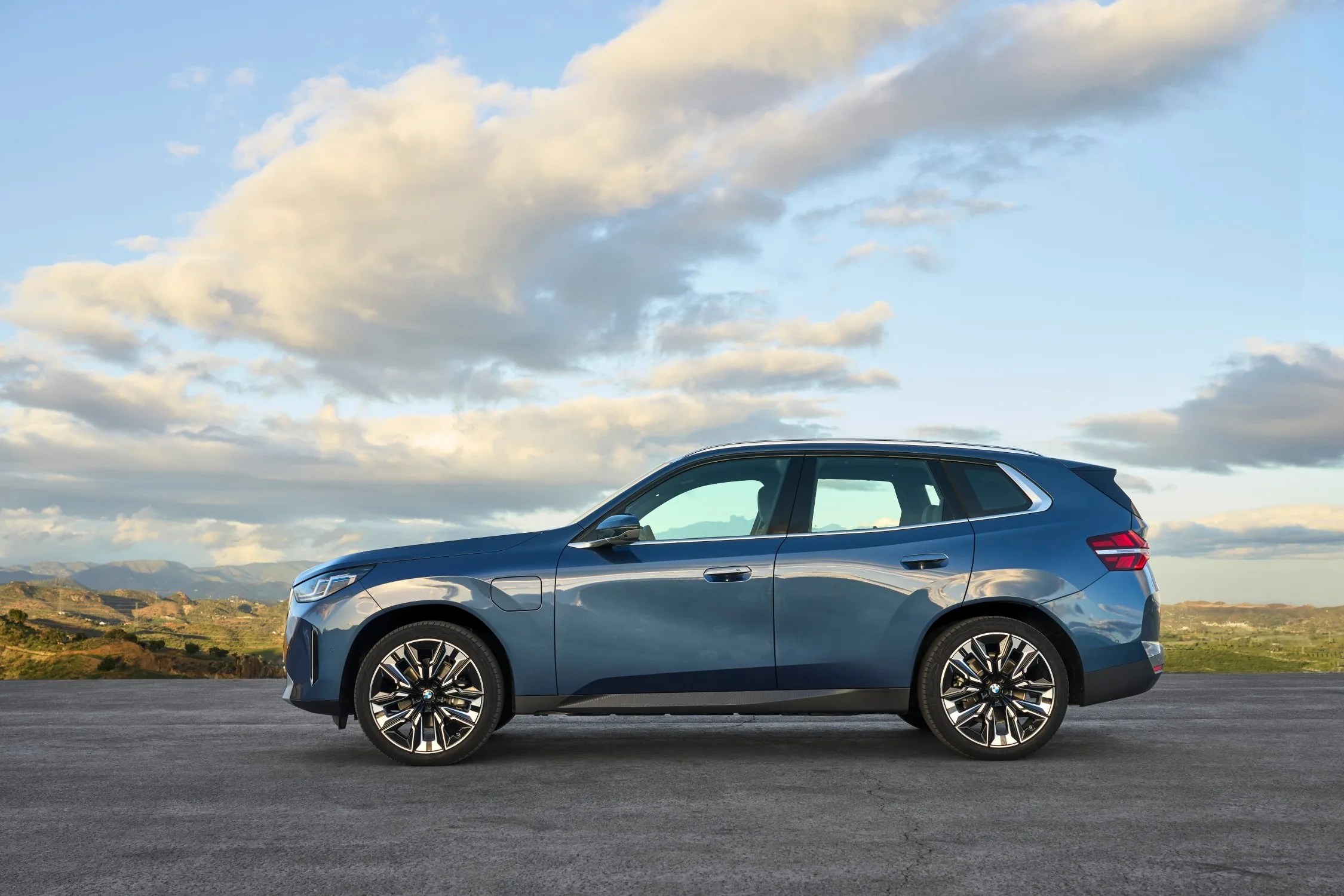 The 2025 X3 shown in bright blue parked in profile view on a large piece of asphalt with a vast view of mountains and a valley in the background.