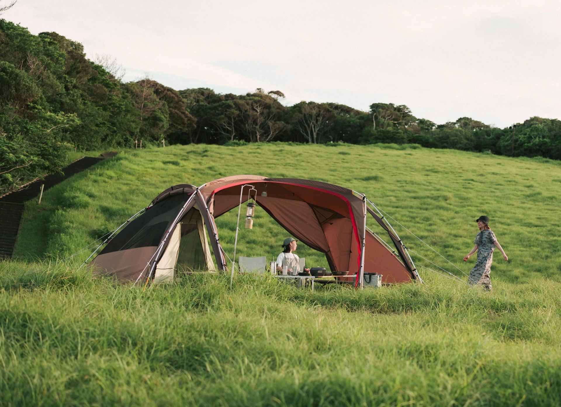 Snowpeak Fubi tent setup in field with woman sitting underneath canopy at table and another woman walking towards the outdoor shelter.