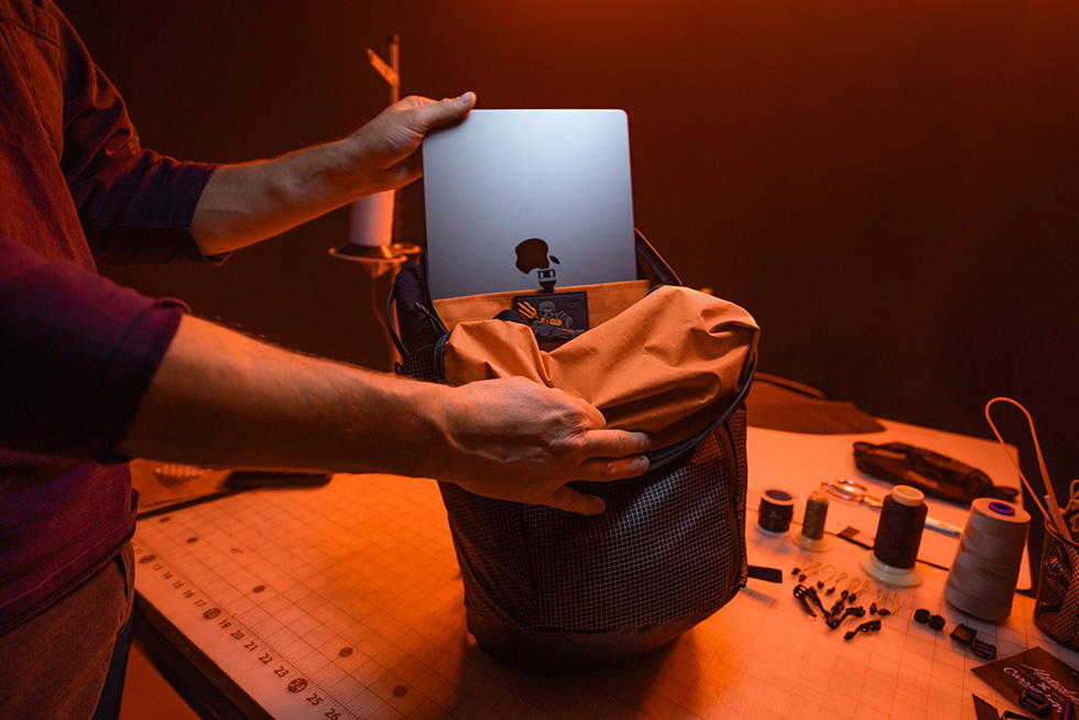 Matador x Carrology EDX Lite backpack opened to reveal a rear tech pocket with an Apple Laptop being slid in. The room appears to be a work table illuminated in an orange hue. 