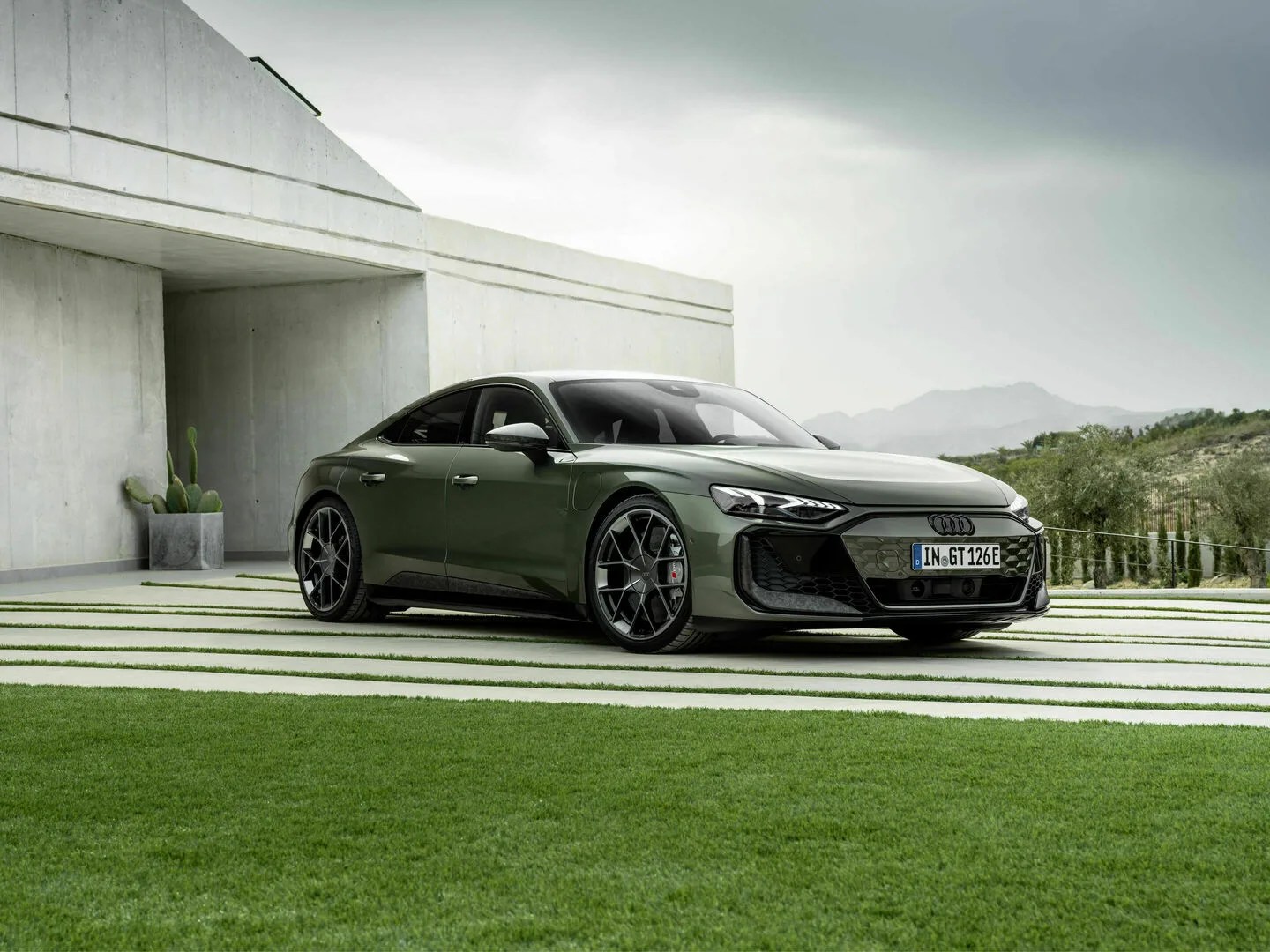 The Audi RS e-tron GT Performance edition parked at an angle on a drive made of strips of concrete with grass lines in between. The car is a deep green color and parked in front of a modern looking concrete building.