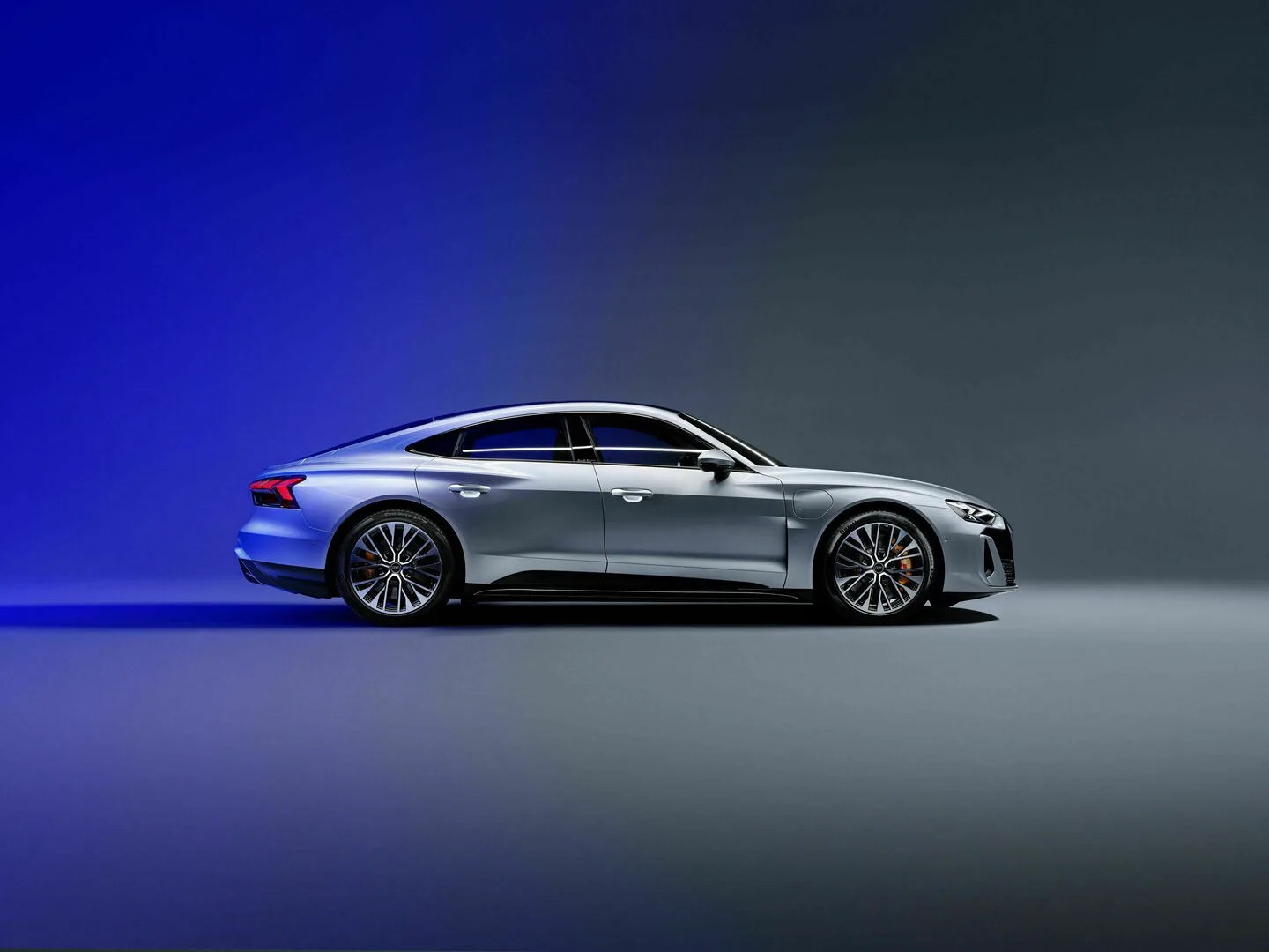 Audi S E-Tron GT shown at a profile view. The car is silver in a featureless grey room, half colored in a blue light. 