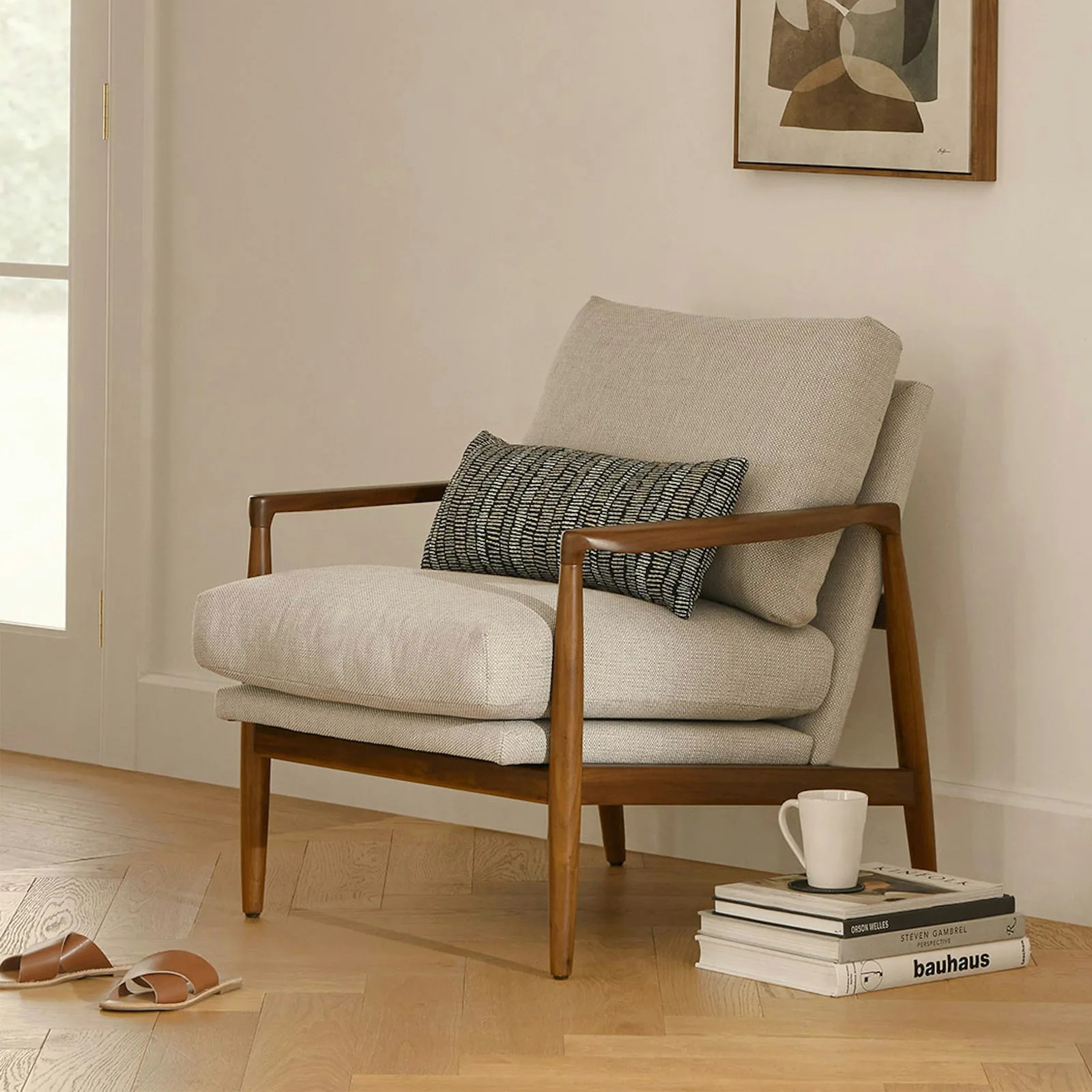 The Article Bavel lounge chair shown pushed against a wall with a pillow resting on the top. A stack of books and a coffee mug sitting on top of the books are pushed against the right hand side of the chair