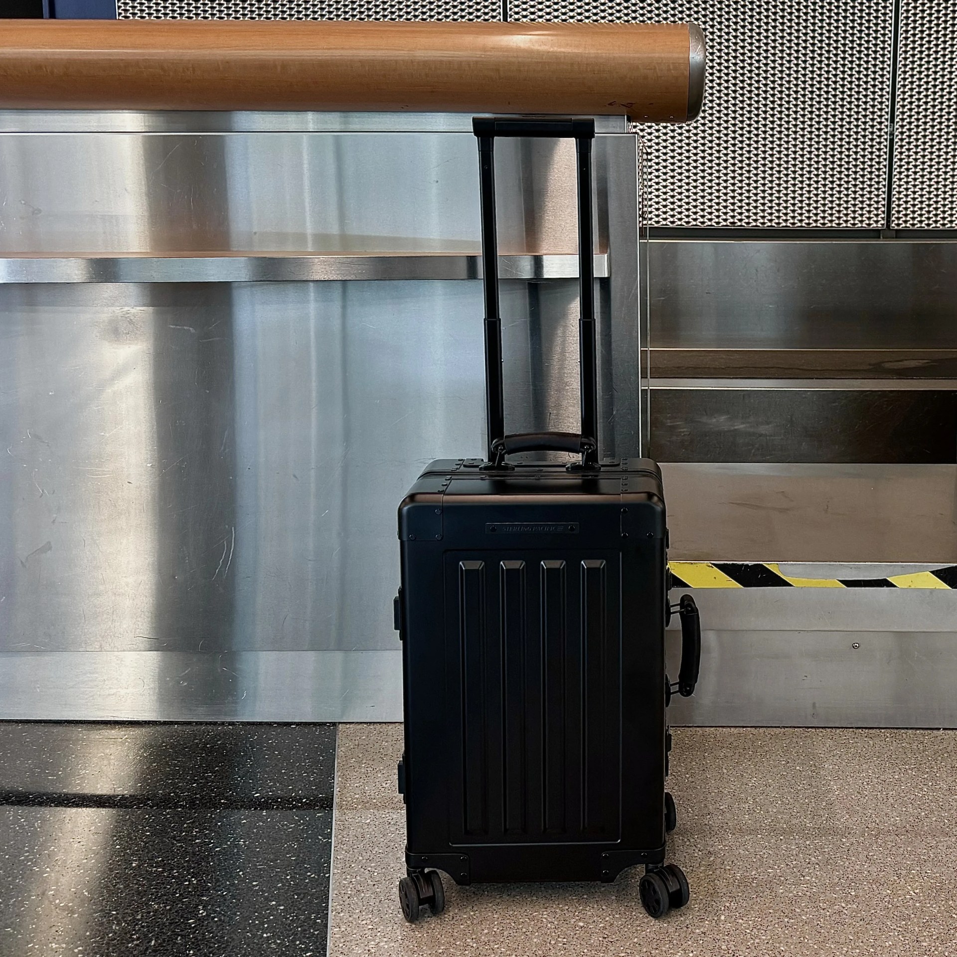 Sterling Pacific 40L Cabin Travel Case aluminum suitcase in black standing in front of an airline check in counter with the handles raised.
