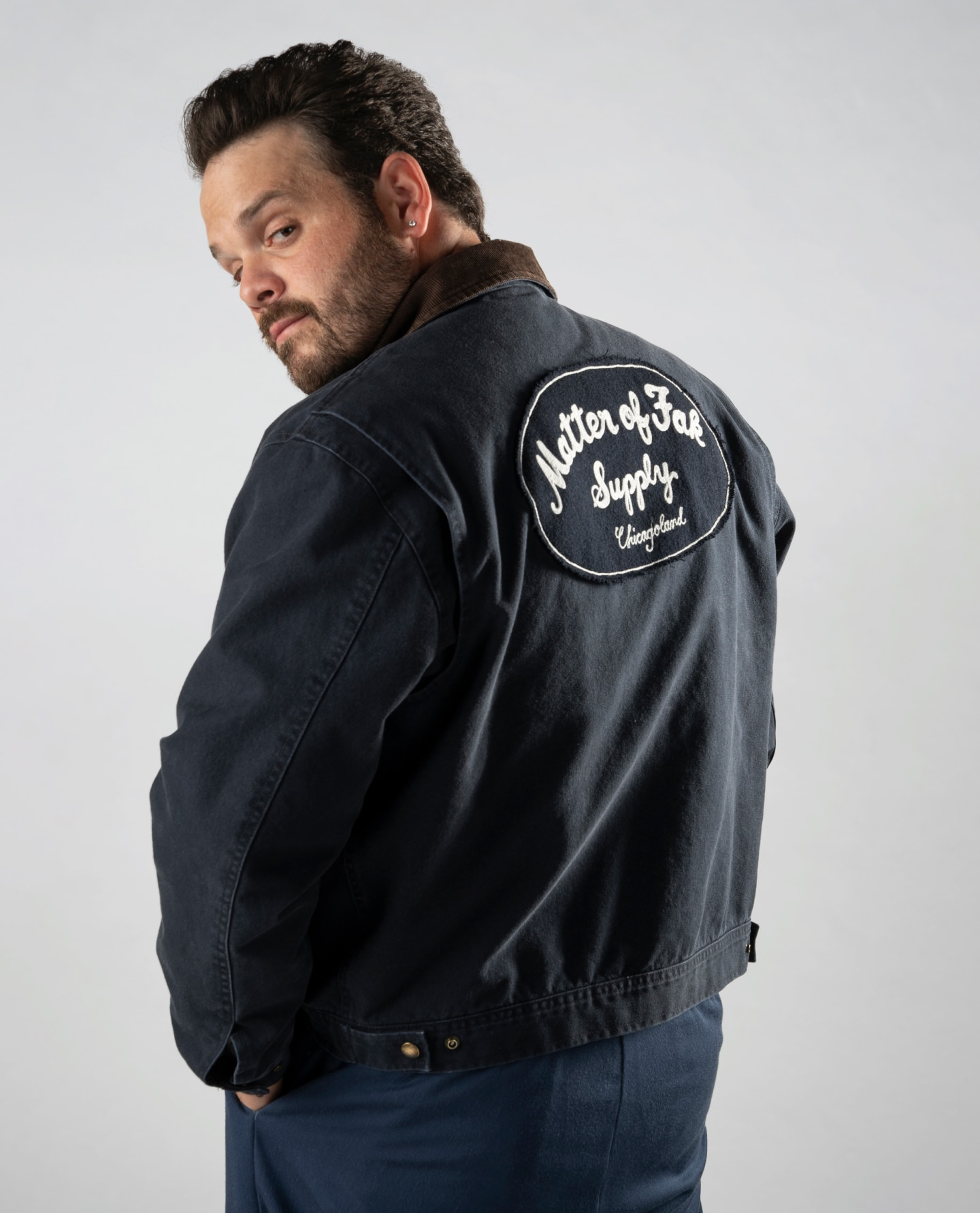 Actor Ricky Staffierni looking back over his left shoulder wearing The Bear x J. Crew collaborative canvas work jacket which shows the Matter of Fak Supply logo on the back.