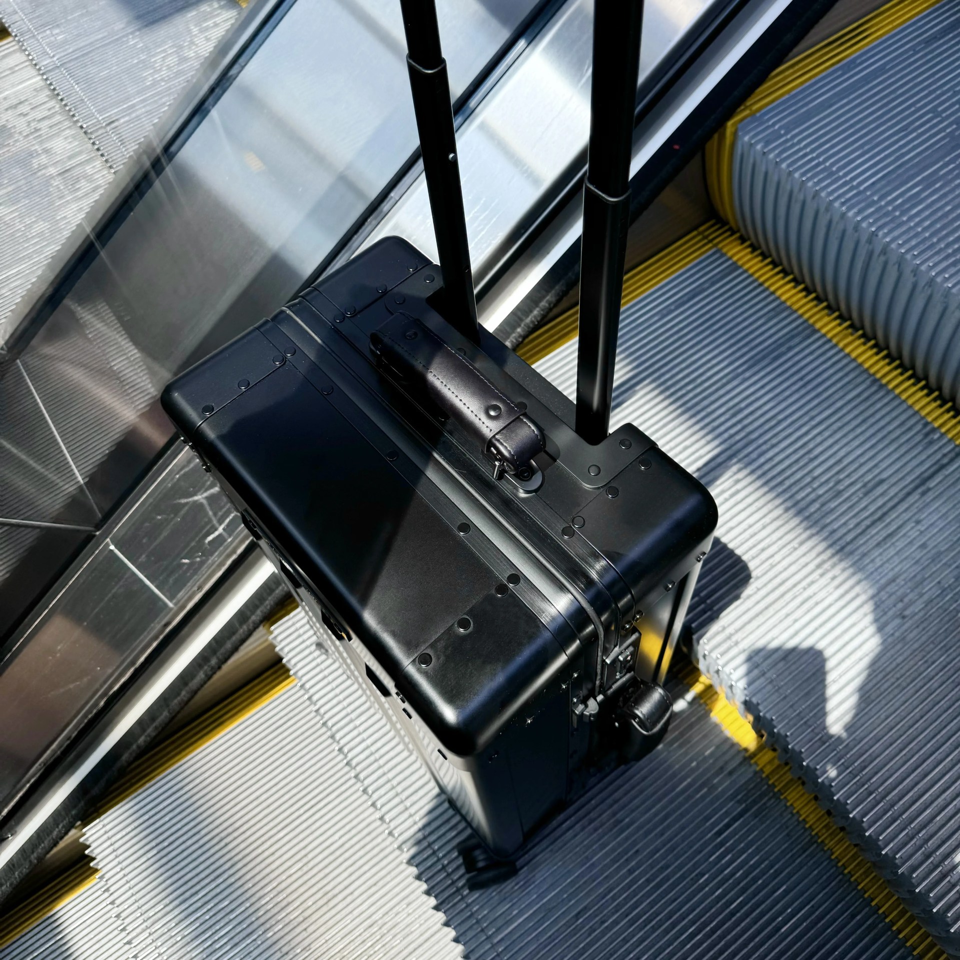 Sterling Pacific 40L Cabin Travel Case in black sitting on an escalator with the handles up. Photo is taken from above looking down at the suitcase. 