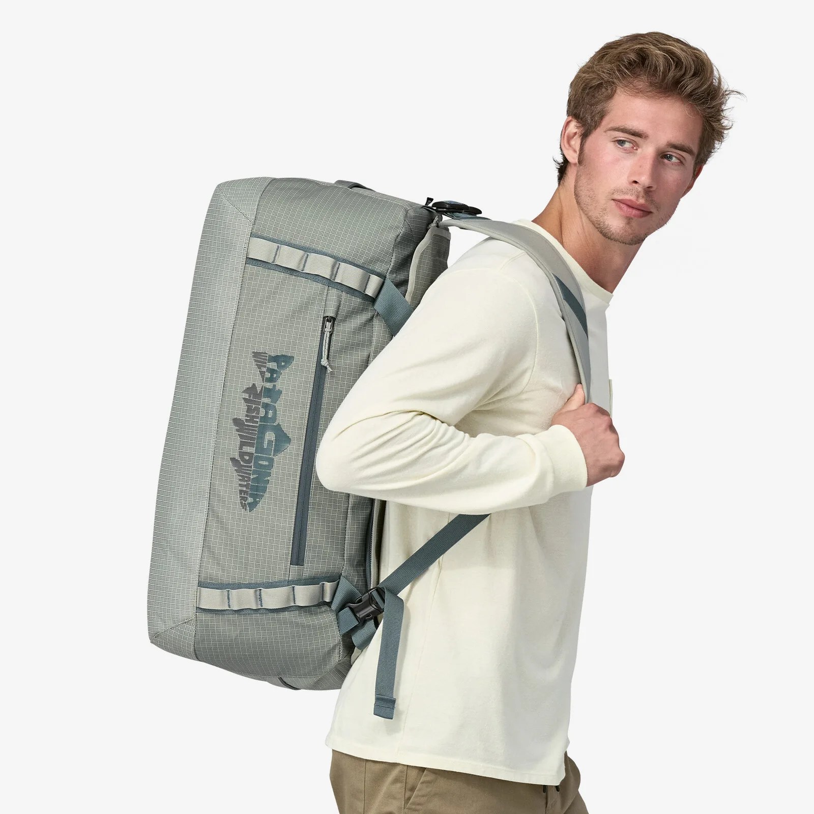A white male carrying a 55l Patagonia Black Hole Duffel on his back like a backpack. He is wearing a cream colored shirt. The bag is a sage green color with a Patagonia fish logo shown on the side.