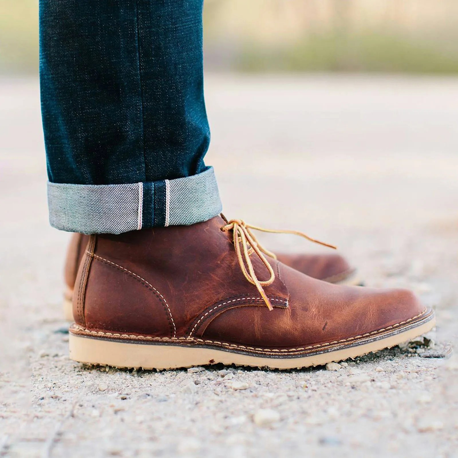 A pair of Red Wing Heritage Weekender Chukkas in reddish brown shown on the feet of a model wearing dark blue jeans rolled at the cuff. The image shows the feet and ankles of the model. The boots are standing on a stoney ground.