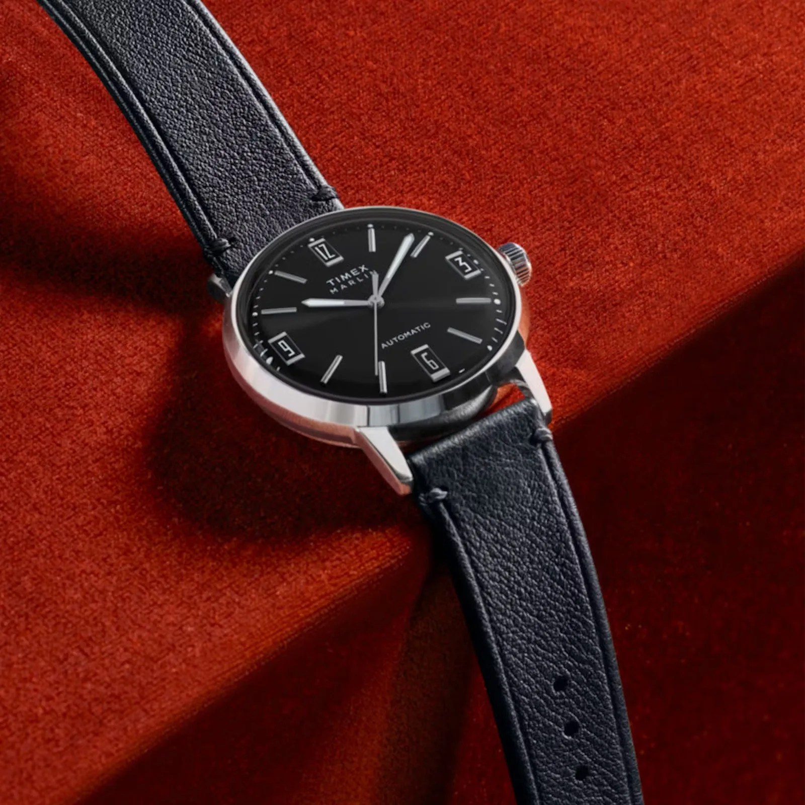 A Timex Marlin Automatic 40mm watch with a black face on a black leather strap lying on a red background.