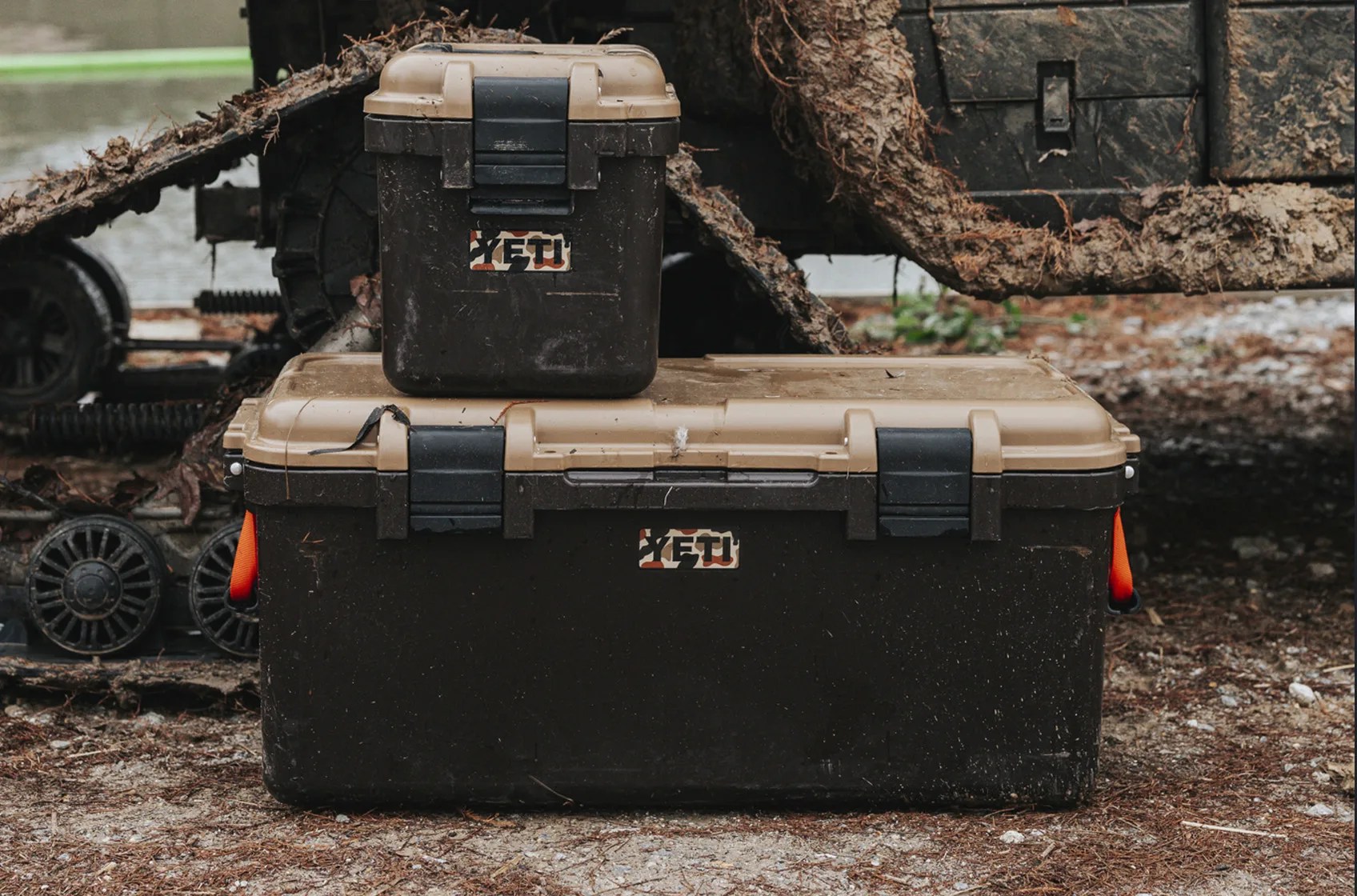 A small Yeti storage box sitting on top of larger yeti storage box in front of a muddy camp ATV. Both Yeti products feature the wetlands color way which is brown and tan with bright orange accents
