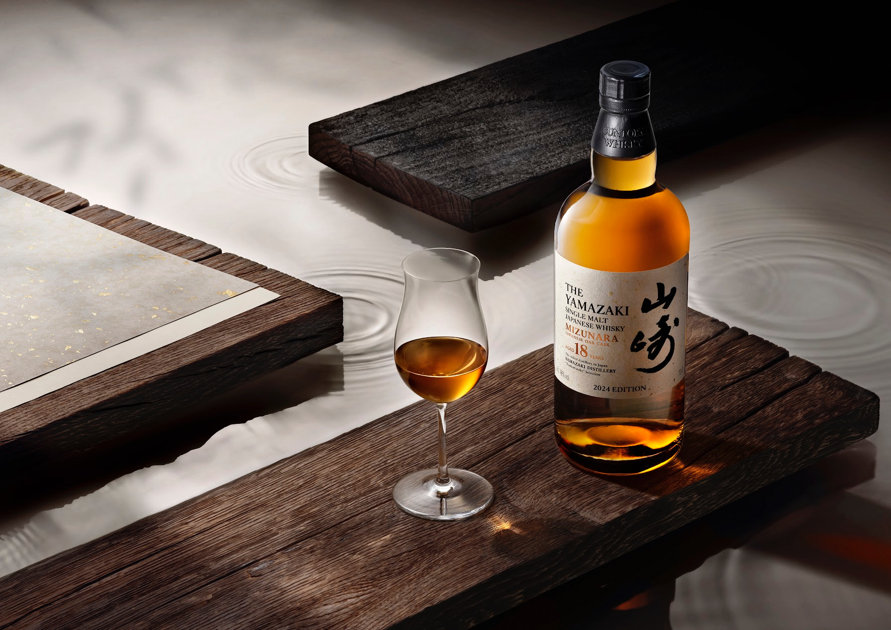 A bottle of The Yamazaki 18-Year-Old Mizunara sitting on a dark plank of wooden next to a glass sitting in the left corner. There’s also a smokey chunk of smoking peat. 