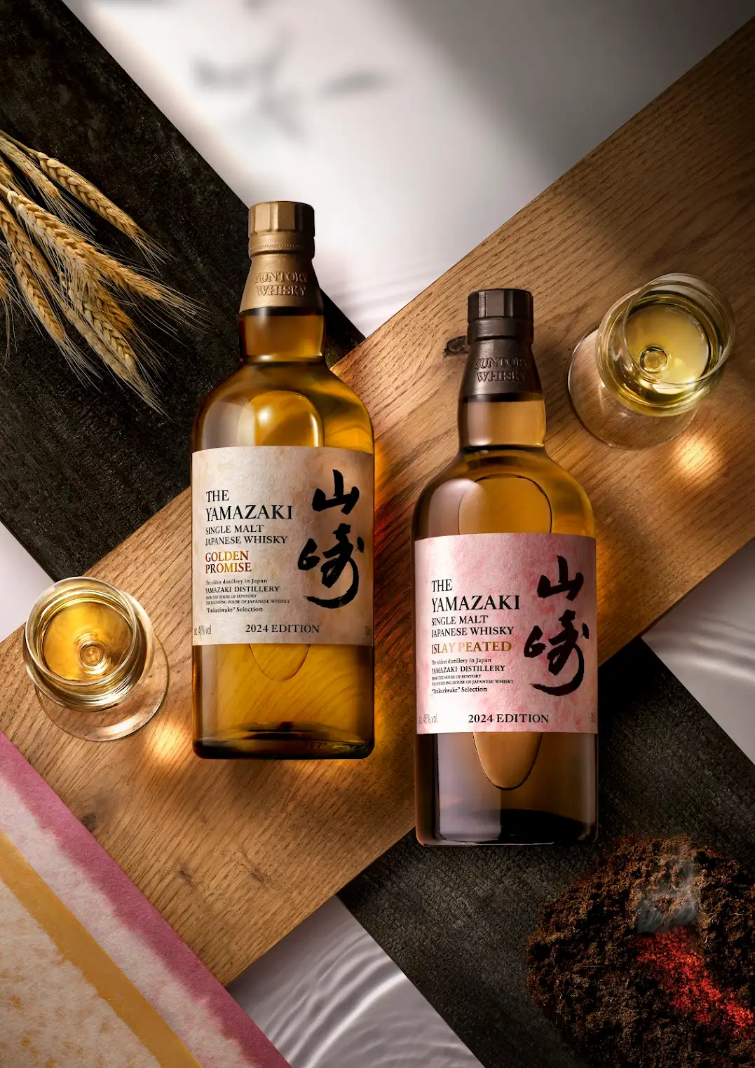 A bottle of The Yamazki Golden Promise and The Yamazaki Islay Peated resting flat on a wooden serving tray flanked by two glasses of whiskey. 