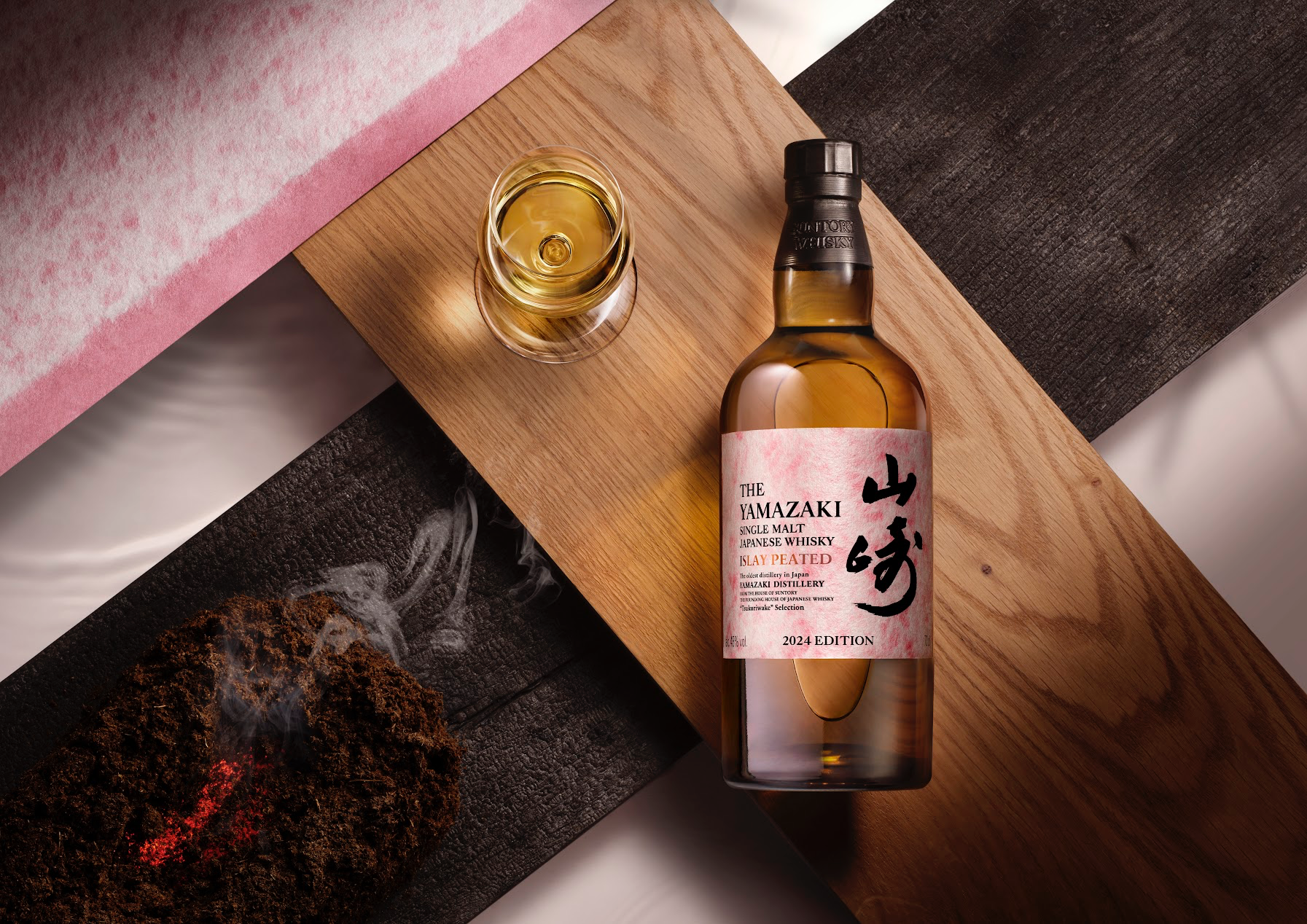 A bottle of The Yamazaki Islay Peated resting flat on a wooden serving tray flanked by a glass of whiskey in the upper left corner. There’s also a smokey chunk of smoking peat. 