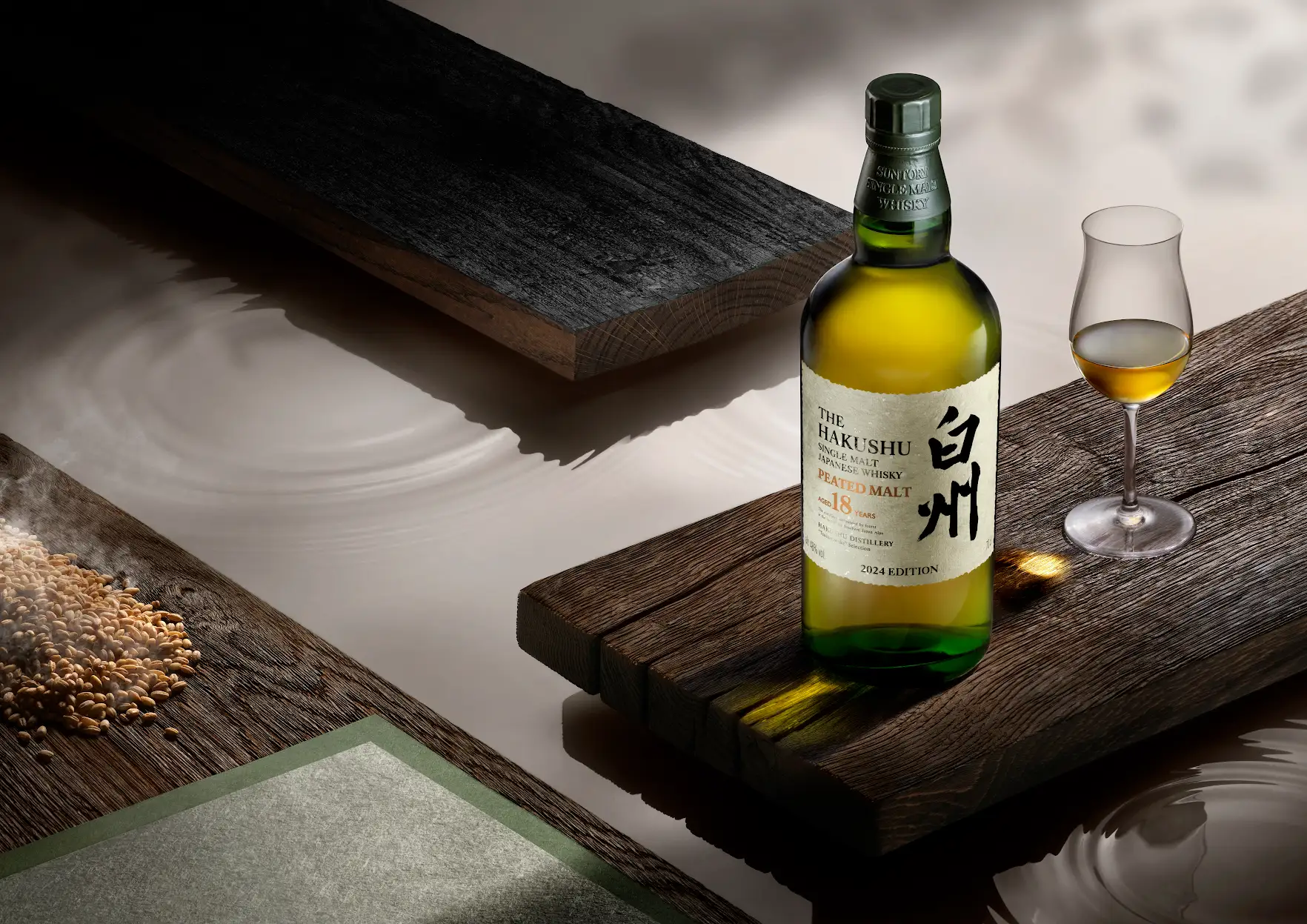 A bottle of The Hakushu 18-Year-Old Peated Malt sitting on a dark plank of wooden next to a glass of whiskey and a pile of peated malted barley seeds.