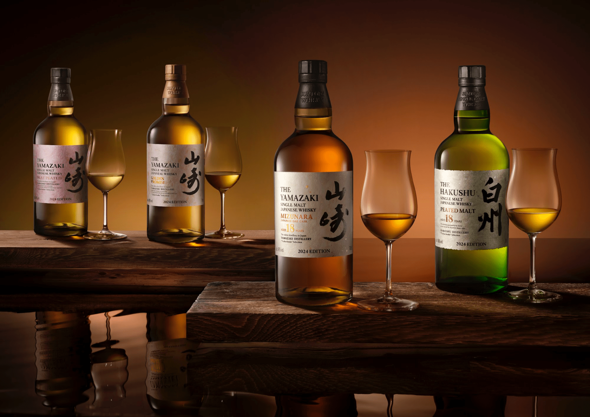 The four bottles included in the 2024 House of Suntory Tsukuriwake collection. Two appear closer sitting on a wood plank, each with a glass of whiskey to the right of the bottle. Two more appear further away in the background also sitting on a wooden plank with a paired glass of whiskey on the right.
