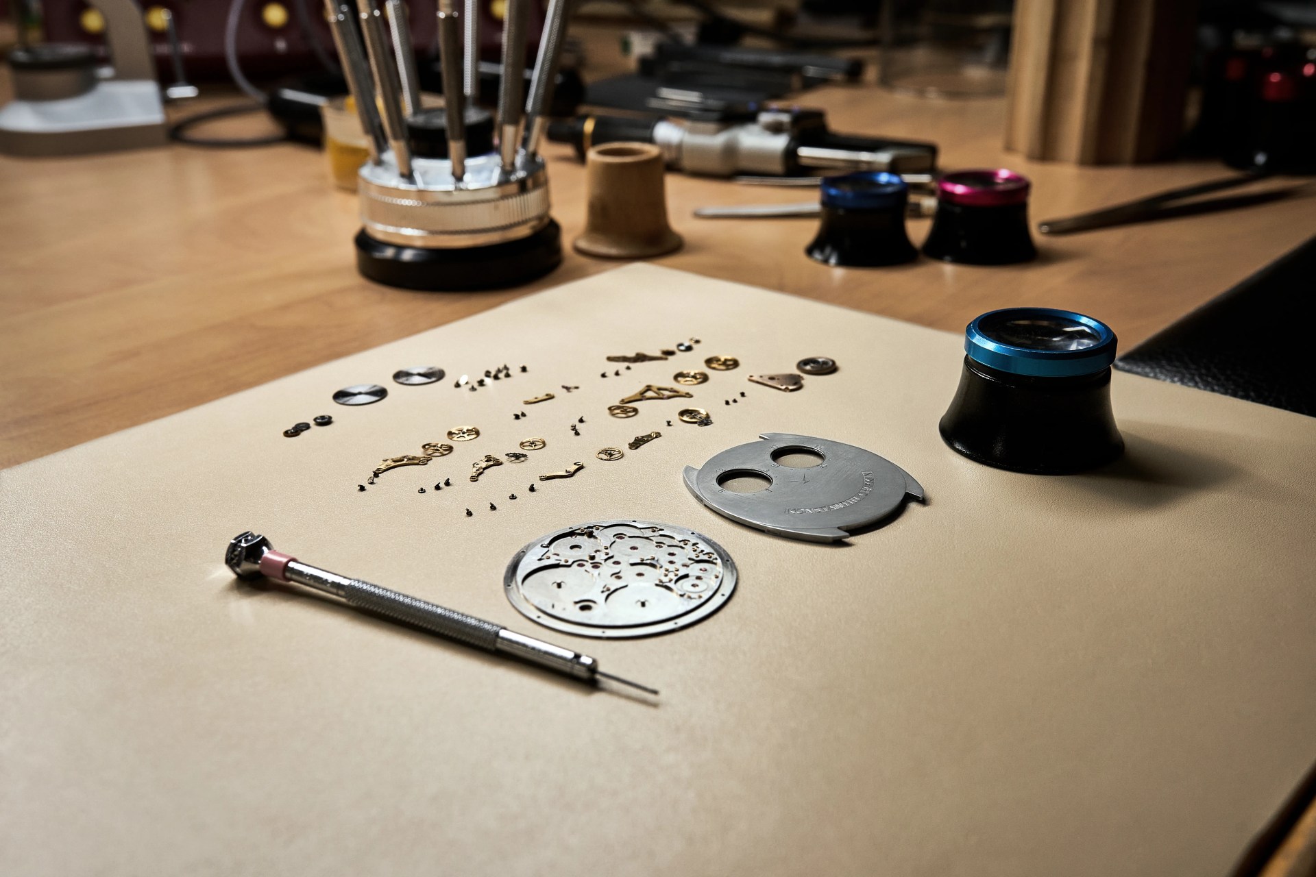 An imagine of the various parts and components of the Konstantin Chaykin ThinKing watch separated and arrange sitting on a table surface next to a watch tool and loupe.